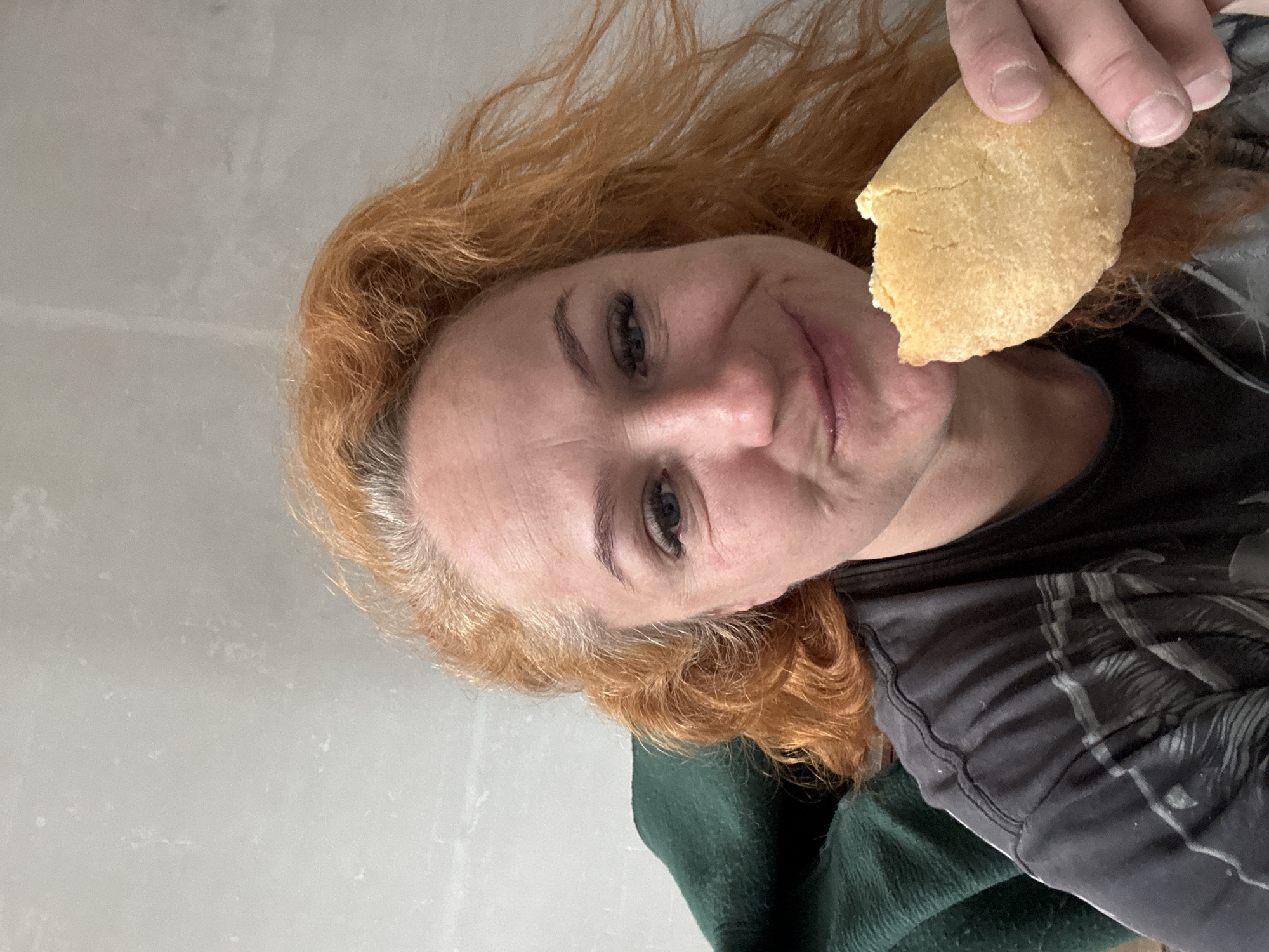 A red-haired woman is holding a partly-eaten round cookie, presumably in Chaikovsky, Russia. She is wearing a dark gray t-shirt with a pattern and a green garment, potentially a sweater or blanket. She has fair skin and blue eyes. The background appears to be a white wall. The image is slightly tilted.