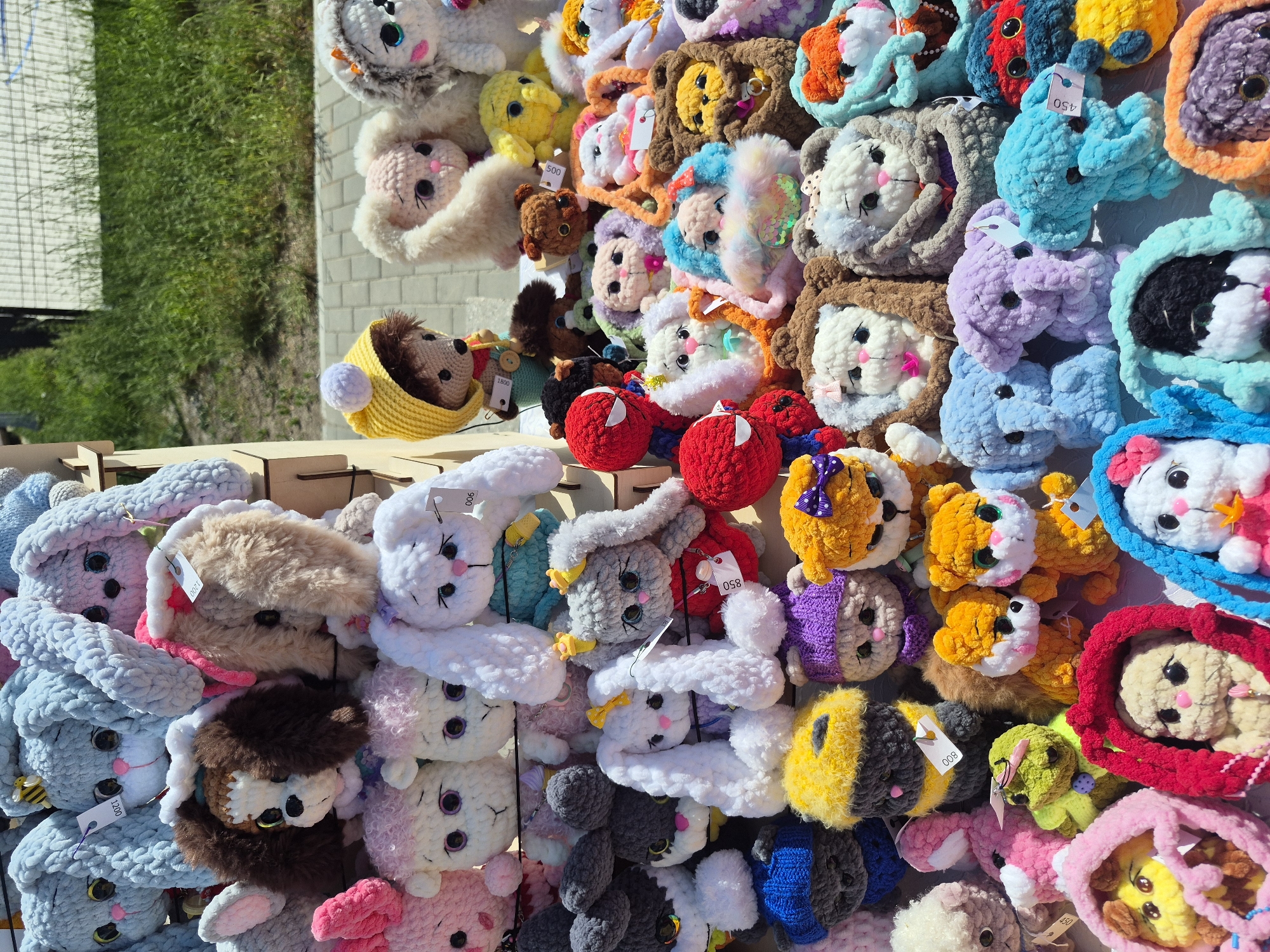 A vendor's outdoor booth displays an array of handmade, plush toys, likely for sale at a market or fair. The toys, mostly depicting animals such as bunnies and lions, are densely packed together and each has a price tag. The environment consists of a grassy area and what appear to be white brick structures in the background. The city and region are unknown.