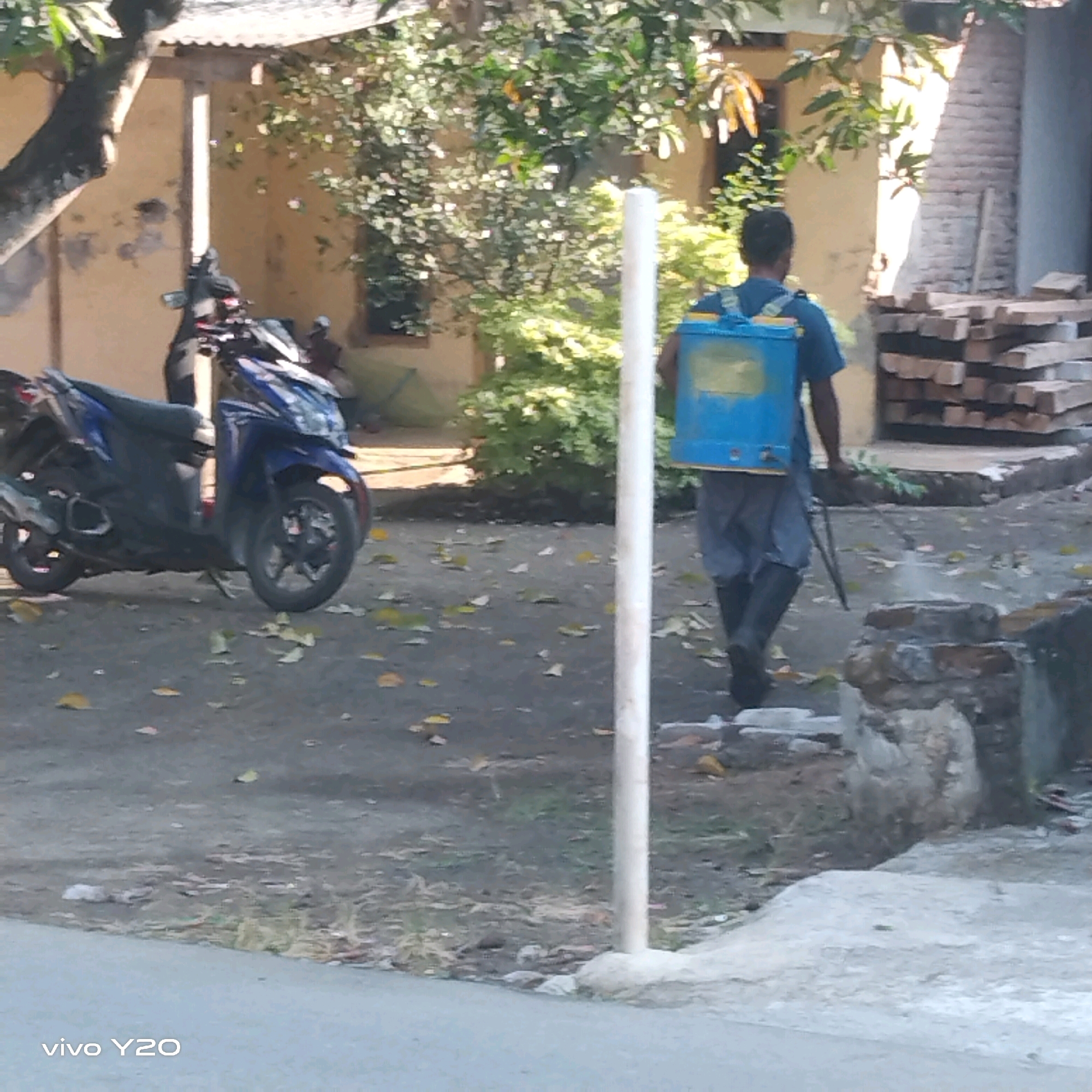 The image shows a person spraying a substance from a blue backpack sprayer. The person is wearing a blue t-shirt and dark pants, and appears to be walking away from the camera. The background includes a blue motorcycle parked on the left, a building with a corrugated metal roof, and a pile of wooden planks on the right. The ground is covered with dirt and fallen leaves. The presence of the motorcycle, building, and overall outdoor setting suggests a scene in a residential or semi-rural area, possibly Karawang, Indonesia, given the context. The action of spraying could be for pest control or agricultural purposes. The lighting indicates it is daytime, with sunlight casting shadows.