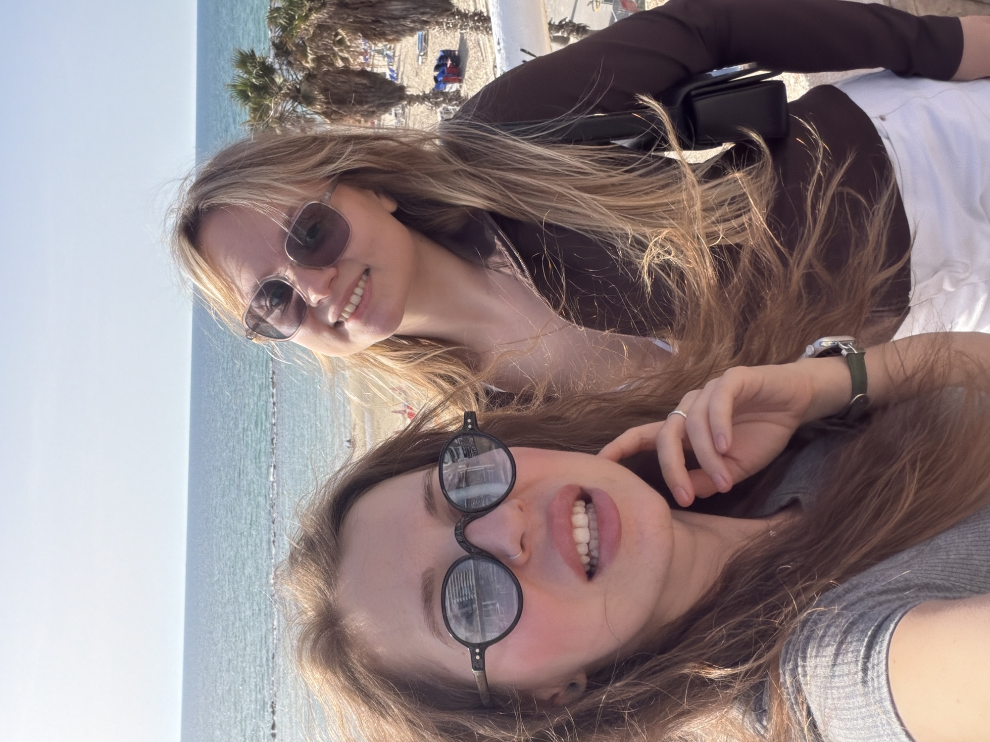 Two young women are posing for a selfie, likely in Bat Yam, Israel, given the presence of a beach and palm trees in the background. The top woman wears sunglasses, a brown top, and white pants, with a small black bag over her shoulder. She is smiling. The bottom woman is closer to the camera, wearing round sunglasses and a grey top. A wristwatch with a green band is visible on her wrist. They are near a beach with a light sky.