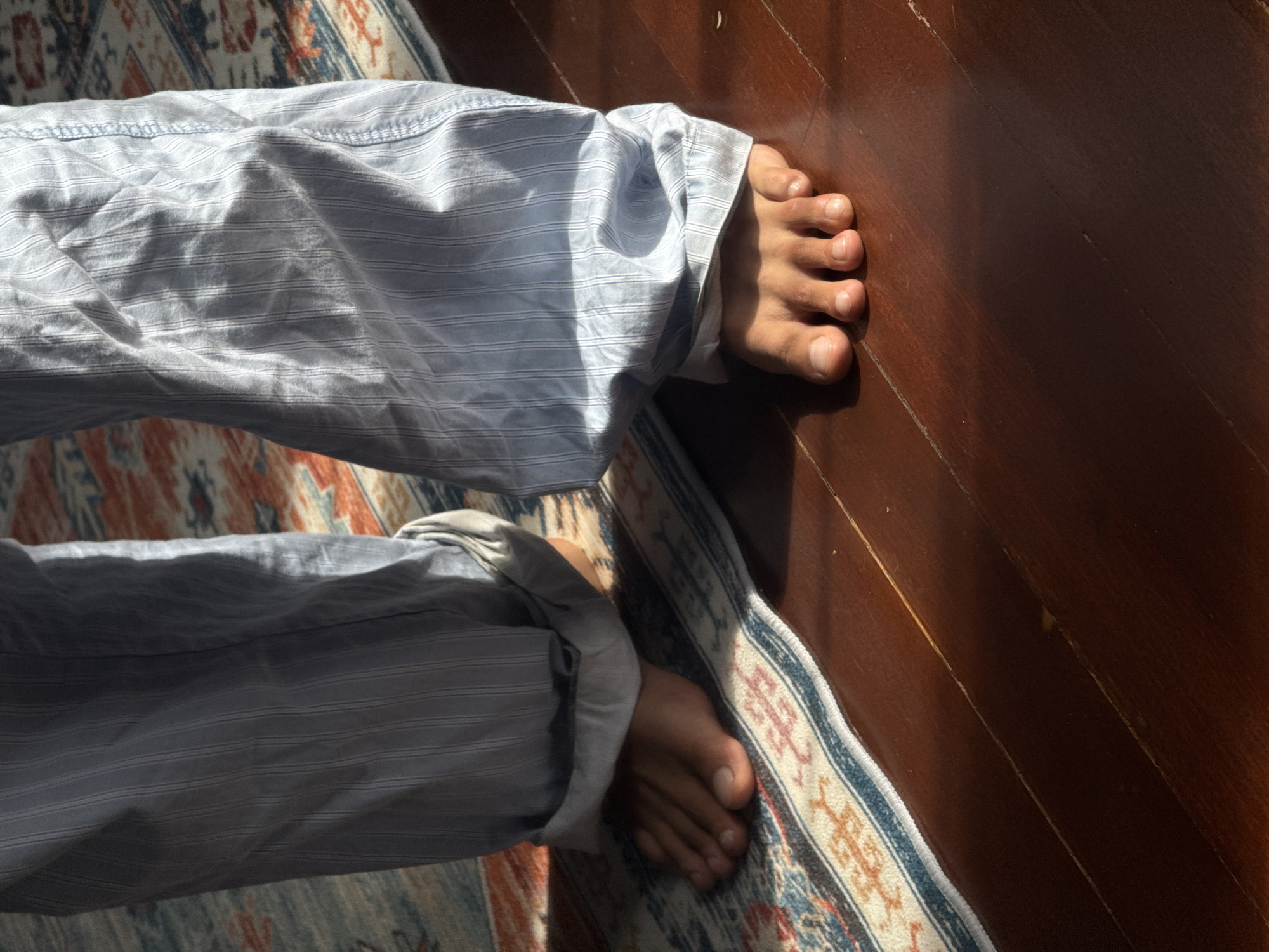 A close-up, low-angle shot focuses on the bare feet of a person standing on a wooden floor partially covered by a patterned rug. The person is wearing light blue striped pajama pants, rolled up at the ankles, exposing their feet. The left foot is on the rug, while the right foot is on the wooden floor. Sunlight casts shadows across the scene, creating a contrast between the illuminated and shadowed areas. The wooden floorboards are visible, and the rug features a geometric pattern in shades of blue, red, and beige. The overall impression is one of domestic comfort and relaxation. The specific location in Kuta Selatan, Indonesia, cannot be determined from the visual information provided.