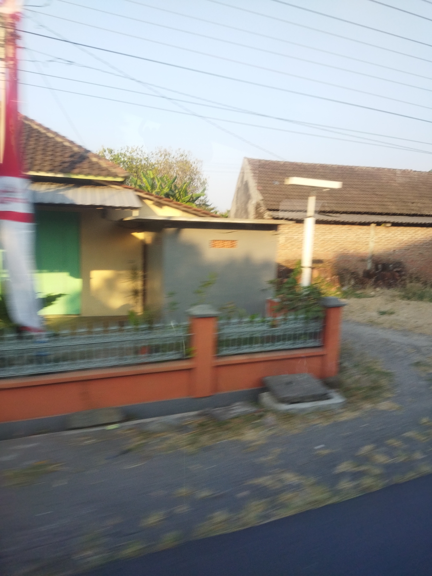 The image captures a blurred view of a street scene in Selogiri, Indonesia, likely from a moving vehicle. In the foreground, a dark asphalt road is visible, with dry grass and debris scattered on its surface. A low, orange-colored wall runs along the edge of the property, topped with a metal fence.
Behind the fence, a small building with a light-colored facade and a corrugated metal roof is partially visible. A green panel or door is on the left side of this building, and a red and white banner or flag is draped vertically on its left edge, also blurred by motion. Further back, a second building with a tiled roof and brick walls is seen. A tall, white pole with a horizontal white element at the top stands between the two buildings. The environment appears to be a residential area with some vegetation. The sky is clear and pale blue. The overall impression is one of a casual, everyday view from a vehicle passing through a semi-urban or rural area in Selogiri.