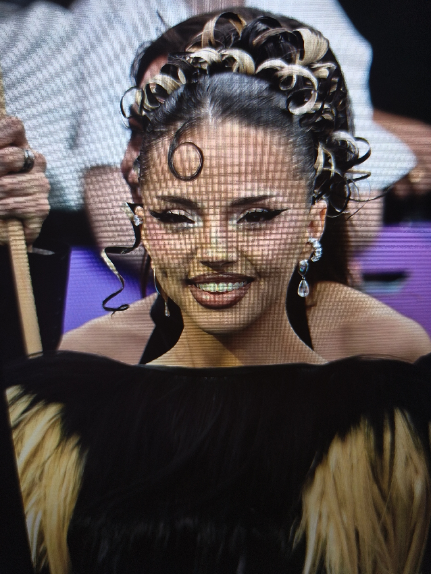 A vibrant close-up image captures a young woman smiling brightly, likely at an indoor event in Svilengrad, Bulgaria. She is the central subject, exuding a confident and joyful emotion.
Her dark hair is styled elaborately, pulled back with numerous sculpted curls, some intricately highlighted with blonde or light brown accents. A distinctive circular curl frames her forehead, and a long, wavy tendril extends near her right temple, adding to her unique look. Her makeup is bold and precise, featuring dramatic winged eyeliner, contoured cheeks with subtle shimmer, and glossy lips. She wears elegant dangling earrings, each adorned with clear, teardrop-shaped stones. Her wide smile reveals her teeth and highlights faint dimples on her cheeks.
She is dressed in a dark garment, possibly a top or dress, from which blonde or light-colored material, resembling fur or feathers, emerges around her shoulders and chest area, visible at the bottom of the frame. This distinctive element contributes to her striking appearance.
In the blurred background, elements suggest a public or formal setting. Parts of other individuals in light-colored attire are visible, along with a prominent purple or violet surface, indicating an event space such as a stage or audience area. To the left, a hand wearing a silver ring is seen holding a wooden stick or pole. The lighting in the image is consistent with an evening event. No discernible text is visible in the media file.