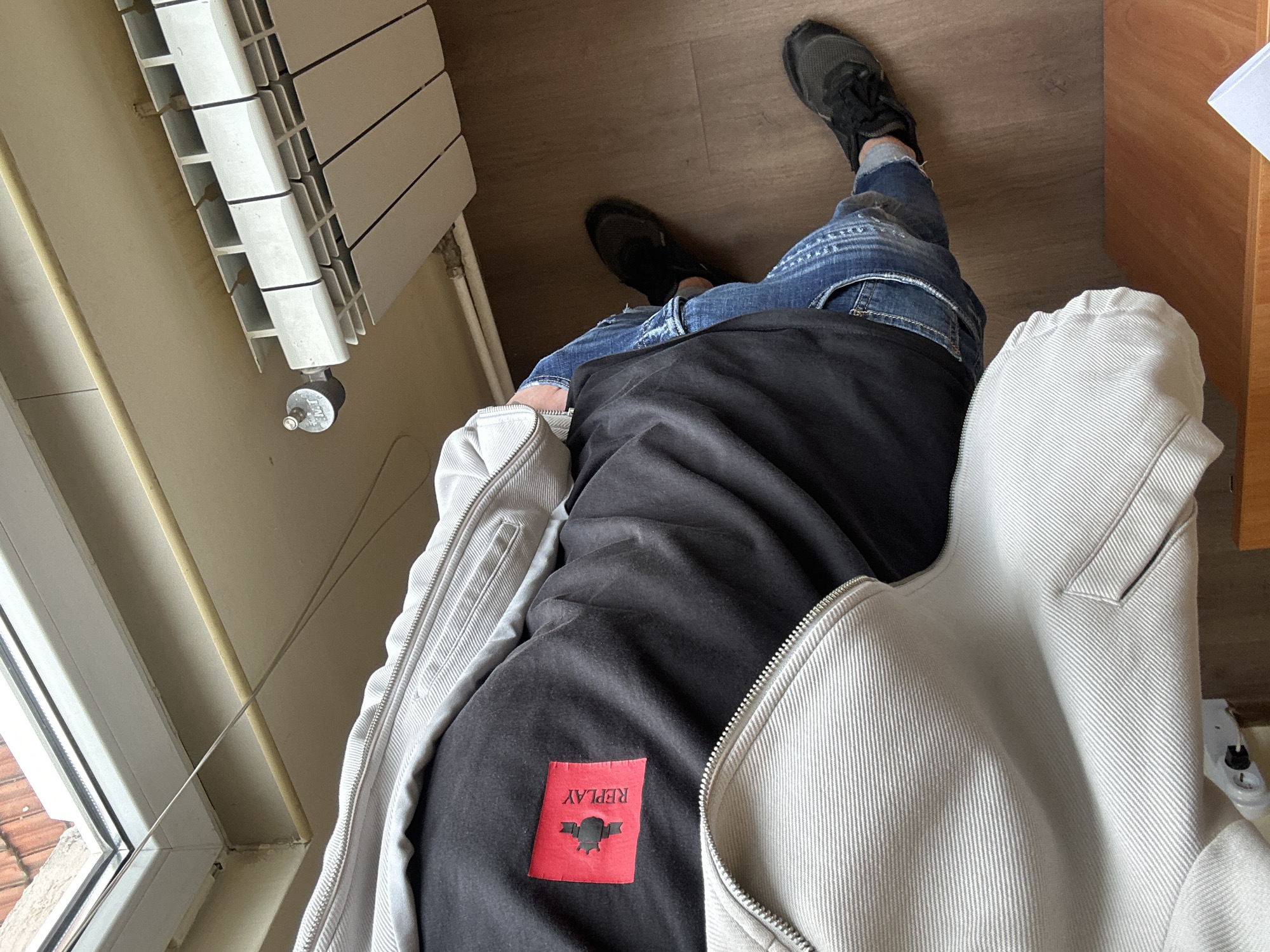 An overhead shot depicts an individual standing indoors, centered on their upper torso and legs. The person wears a light grey zippered jacket made of a ribbed or corduroy-like fabric, partially open to reveal a black long-sleeved shirt underneath. A red rectangular patch with a black stylized logo and the word "REPLAY" is visible on the upper back of the black shirt. Below, the individual wears distressed blue jeans with visible rips and fading, and dark athletic shoes. The flooring is a light brown wood-effect laminate or parquet. In the upper left quadrant, a white-framed window looks out onto a terracotta-tiled roof, with a thin wire running horizontally near the window frame. To the right, a white multi-sectioned panel radiator with a metallic valve, marked "M.T.", is mounted on a light beige wall. A thin wooden dowel or rod runs horizontally along the wall above the radiator. The scene is located within a room in Plovdiv, Bulgaria.