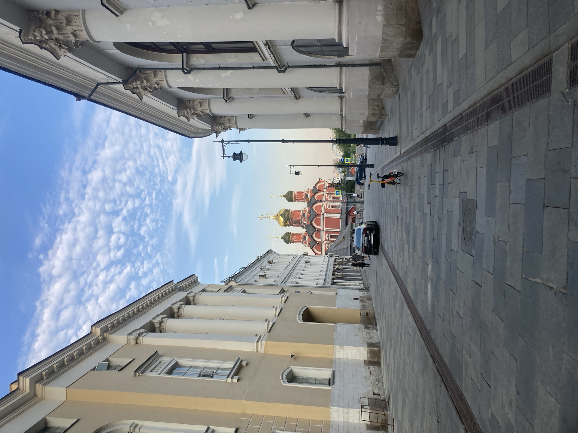 This is a vertical, medium-shot, eye-level photograph taken from a street in Moscow, Russia, looking towards a large red building with golden domes, identified as the Cathedral of the Assumption of the Blessed Virgin Mary in the Kremlin. The foreground of the image features a wide, paved street with large, gray stone tiles. A metal grate runs along the center of the street, suggesting a drainage system. On the right side of the street, a building with white, classical architecture is visible, characterized by large, fluted columns and arched windows. On the left side, a building with lighter yellow-colored facade is seen, also with classical architectural elements like columns and rectangular windows.
In the middle ground, several elements are present: a black sedan car is parked on the right side of the street, and two bicycles, one orange and one yellow, are parked in front of it. A streetlamp with a black pole and a lamp fixture is positioned on the right side of the street, and another similar lamp is visible further down the street. People are walking on the sidewalk on the left side of the street. The background is dominated by the red cathedral with its prominent golden domes, standing against a bright blue sky filled with scattered, fluffy white clouds. The overall atmosphere of the image is serene and picturesque, capturing a blend of historical architecture and urban life.