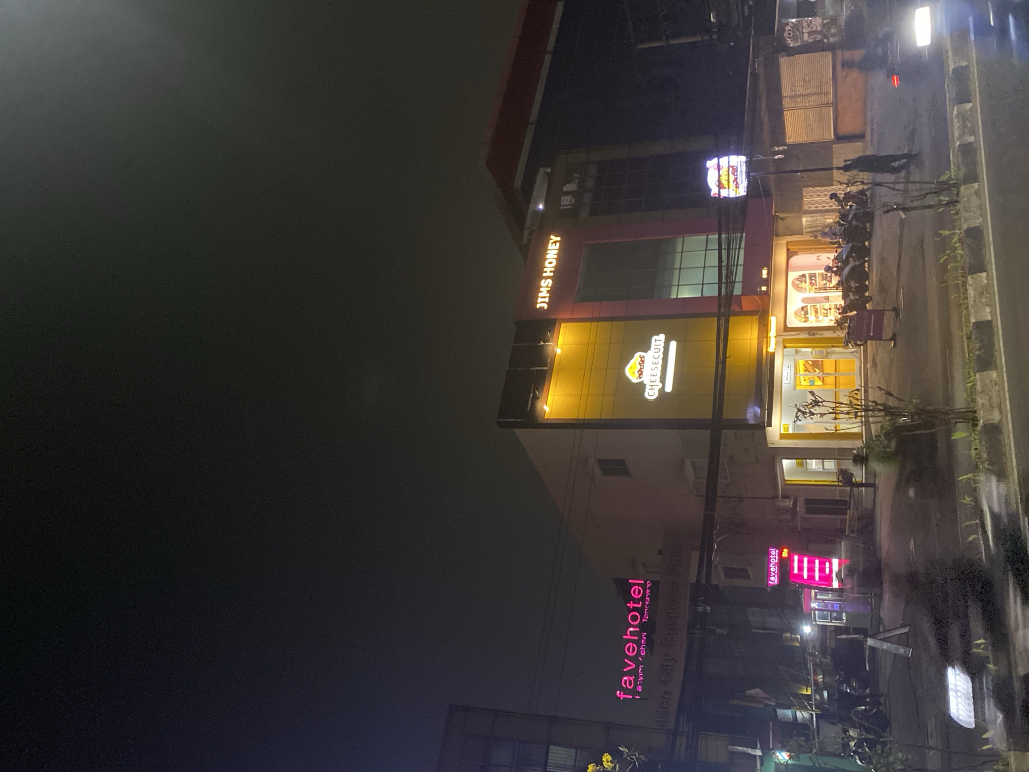 The image captures a nighttime scene on a street in Tangerang, Indonesia. The focal point is a building with illuminated signage. On the left, a pink neon sign reads "favehotel" with smaller text below. Further right, a yellow building features a sign with a cartoon cheese character and the text "CHEESECUIT." Above these, a larger sign displays "JIMS HONEY" in illuminated letters. The street in front of the buildings is lined with parked motorcycles. A person can be seen walking on the sidewalk on the right side of the frame, and the headlights of a vehicle are visible in the foreground. The sky above is dark and appears overcast or hazy.
