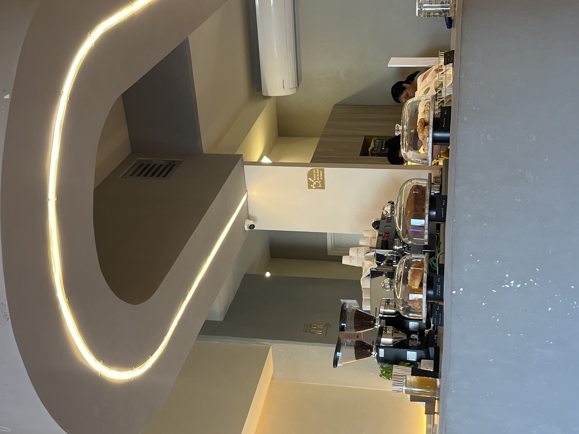 This image captures the interior of a modern coffee shop or cafe, characterized by clean lines and neutral tones. A prominent feature is a long service counter that extends across the mid-ground, equipped with multiple coffee grinders, espresso machines, and stacked cups, some labeled "CAFÉS."
Displayed on the counter under clear glass domes are various pastries, including "GALLETA SAL" (salty cookie), "CROISSANT," and "PANQUE" (pound cake), available for purchase. Behind the counter, a person with dark hair, likely a barista or staff member, is partially visible, attending to the area.
The cafe's design includes an architectural curve in the upper left of the frame, illuminated by a warm-toned LED strip light. Walls are painted in shades of grey and beige, and an air conditioning unit is mounted high on a wall. A security camera (CCTV) is visible on a wall further back.
Notable text includes a yellow sign on a wall stating, "APROBADO 100% PARA SER VIRAL Y TE ENCANTE" (Approved 100% to be viral and you love it), alongside an icon. Another sign shows two gender symbols with an arrow, possibly indicating restrooms or a specific direction. The scene depicts a functional, well-lit indoor environment, suggesting a typical daytime operation in a cafe setting in San Bernardino Tlaxcalancingo, Mexico.