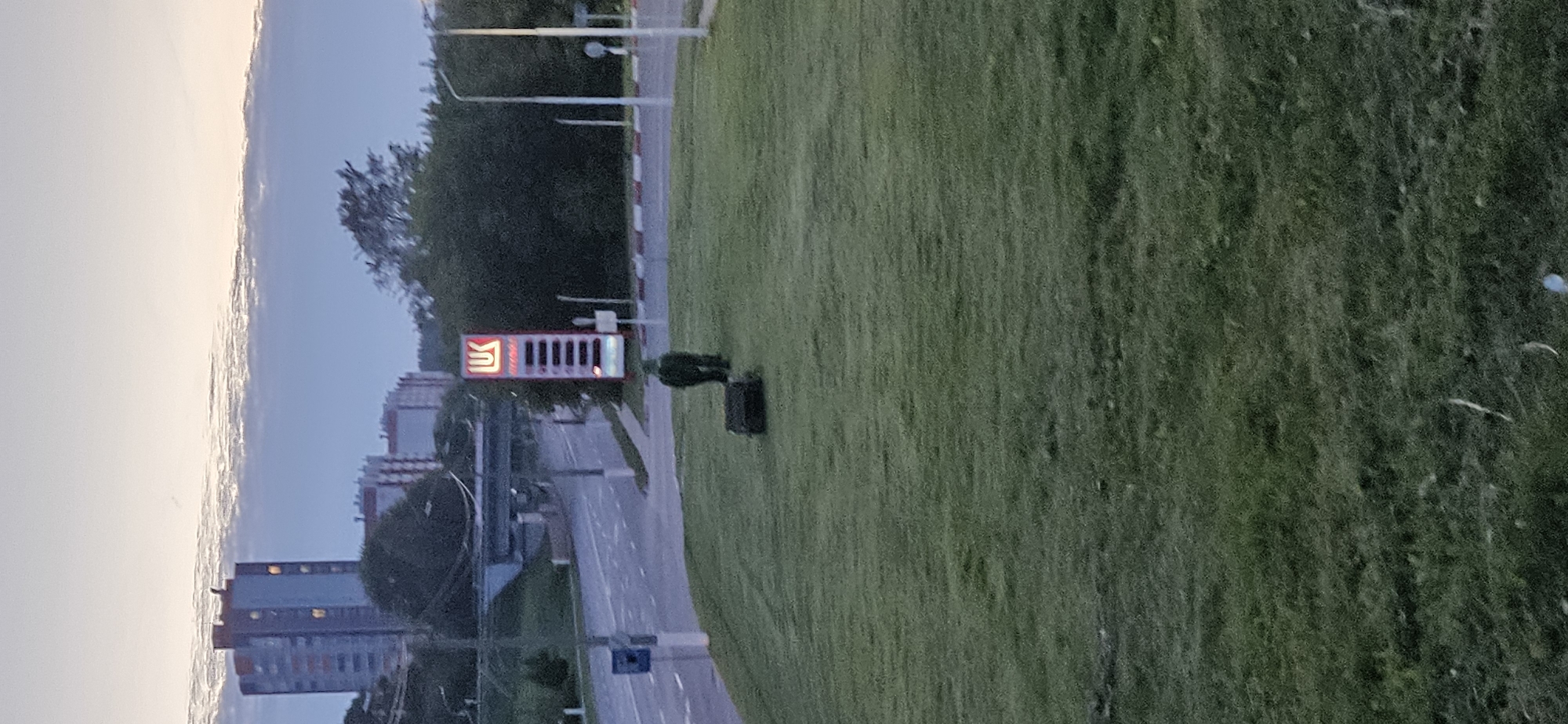 The image depicts an outdoor scene at dusk, likely in Vitebsk, Belarus, given the presence of a Lukoil gas station sign. In the foreground, a grassy hill slopes downwards, with a dark, rectangular object, possibly a trash receptacle or a small structure, situated on the grass. Further up the hill and to the left, a person is visible, appearing to be walking away from the camera and towards the gas station. This individual is wearing dark, possibly camouflage, clothing.
In the mid-ground, a road stretches across the scene, with a visible traffic lane and a pedestrian walkway. A Lukoil gas station tower with illuminated pricing information is prominent on the right side of the road. Beyond the road, a concrete overpass or bridge is visible, and in the background, apartment buildings with illuminated windows suggest an urban environment. The sky above is a soft gradient of pale yellow and blue, with wispy clouds indicating the transition from day to night. The overall atmosphere is serene and quiet, characteristic of early evening in a city.