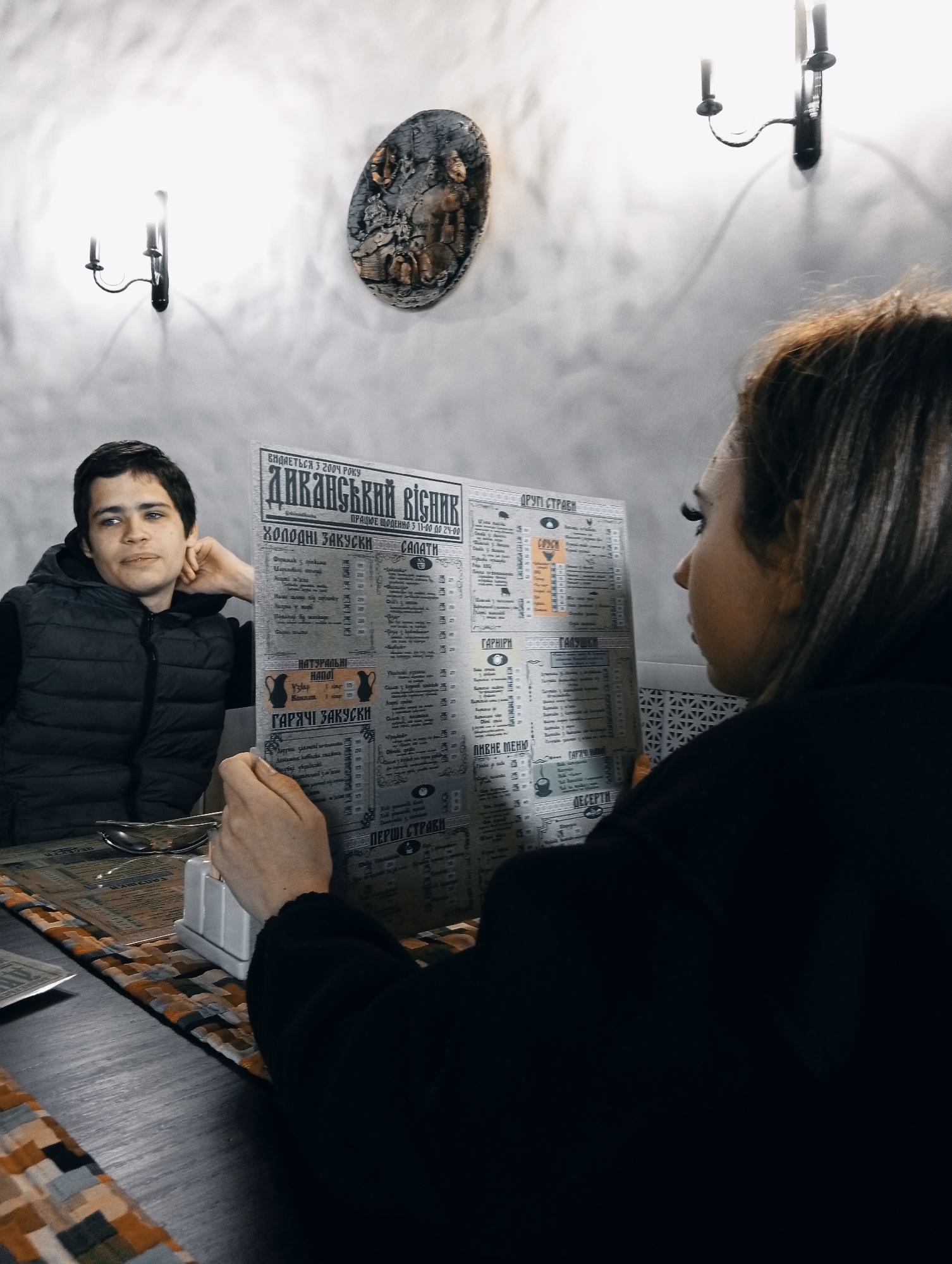 The image shows two people at a table in a restaurant, likely in the city of Poltava, Ukraine, based on the Cyrillic script in the menu. A man in a dark vest sits to the left, resting his head on his hand and looking away from the viewer. A woman in a black jacket sits to the right, holding a menu titled "Диканський вісник", which translates to "Dikansky Bulletin", an actual restaurant name, and looking at it intently. The menu features sections like "Холодні закуски" (Cold snacks), "Салати" (Salads), "Гарячі закуски" (Hot snacks), "Перші страви" (First courses), "Другі Страви" (Second courses), "Гарніри" (Side Dishes), "Гарячі напої" (Hot drinks), "Десерти" (Desserts), and "Пивне меню" (Beer menu).
The table is covered with a patterned placemat, and there's a condiment holder and some cutlery on it. The background features a textured white wall, decorated with sconces and a round decorative plaque.