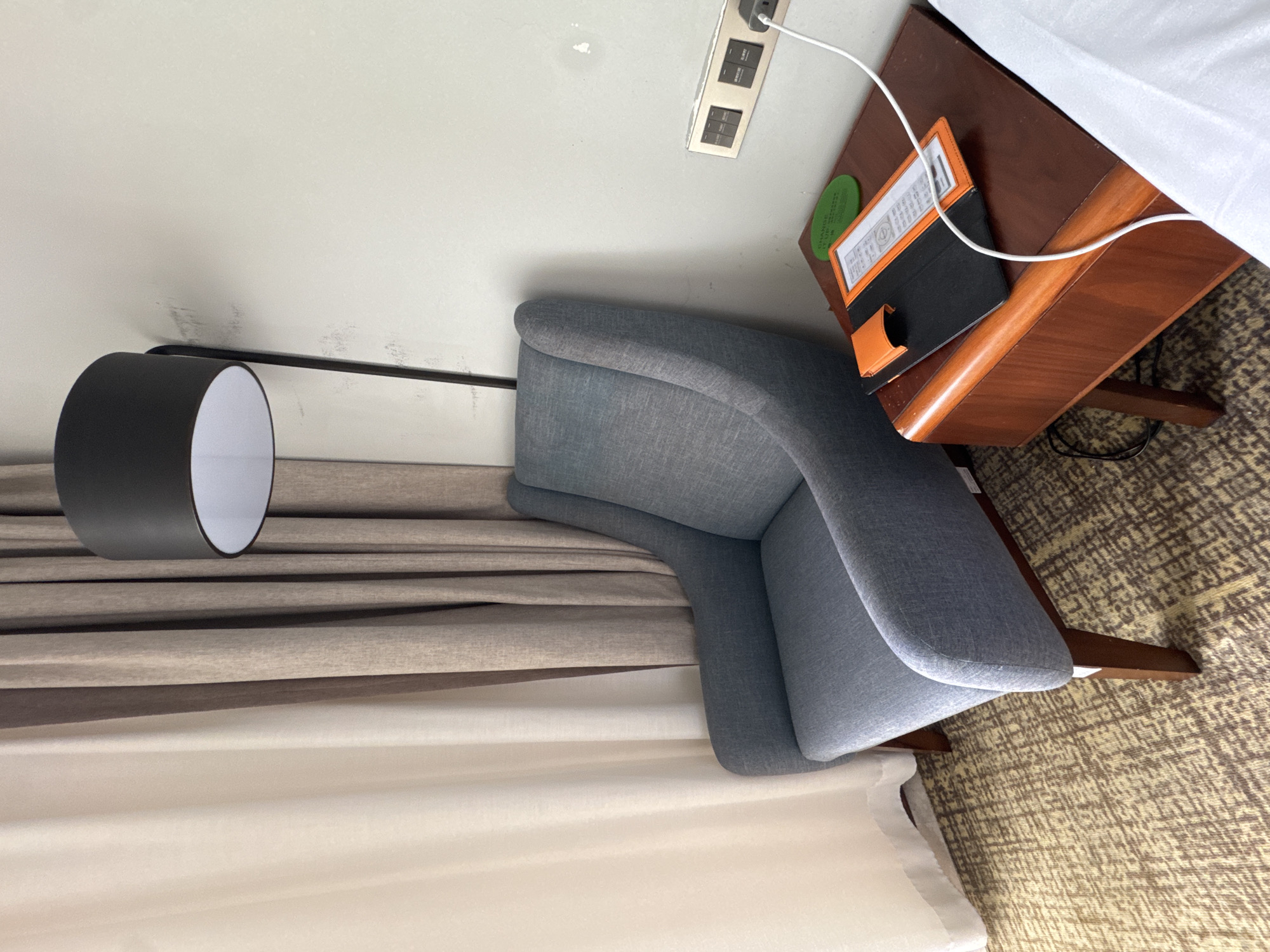 A floor lamp with a black shade stands behind a blue upholstered armchair. The armchair is situated next to a small wooden side table on which a white charging cable is plugged into a wall outlet. On the table, there is a black folder with an orange trim and a green circular object with text. The scene is within a room in Baiyun District, China, indicated by the presence of hotel amenities such as the charging cable and what appears to be a hotel information card or menu on the table, along with a light-colored curtain in the background. The floor is carpeted with a textured brown and tan pattern. The wall is light-colored and shows some minor imperfections.