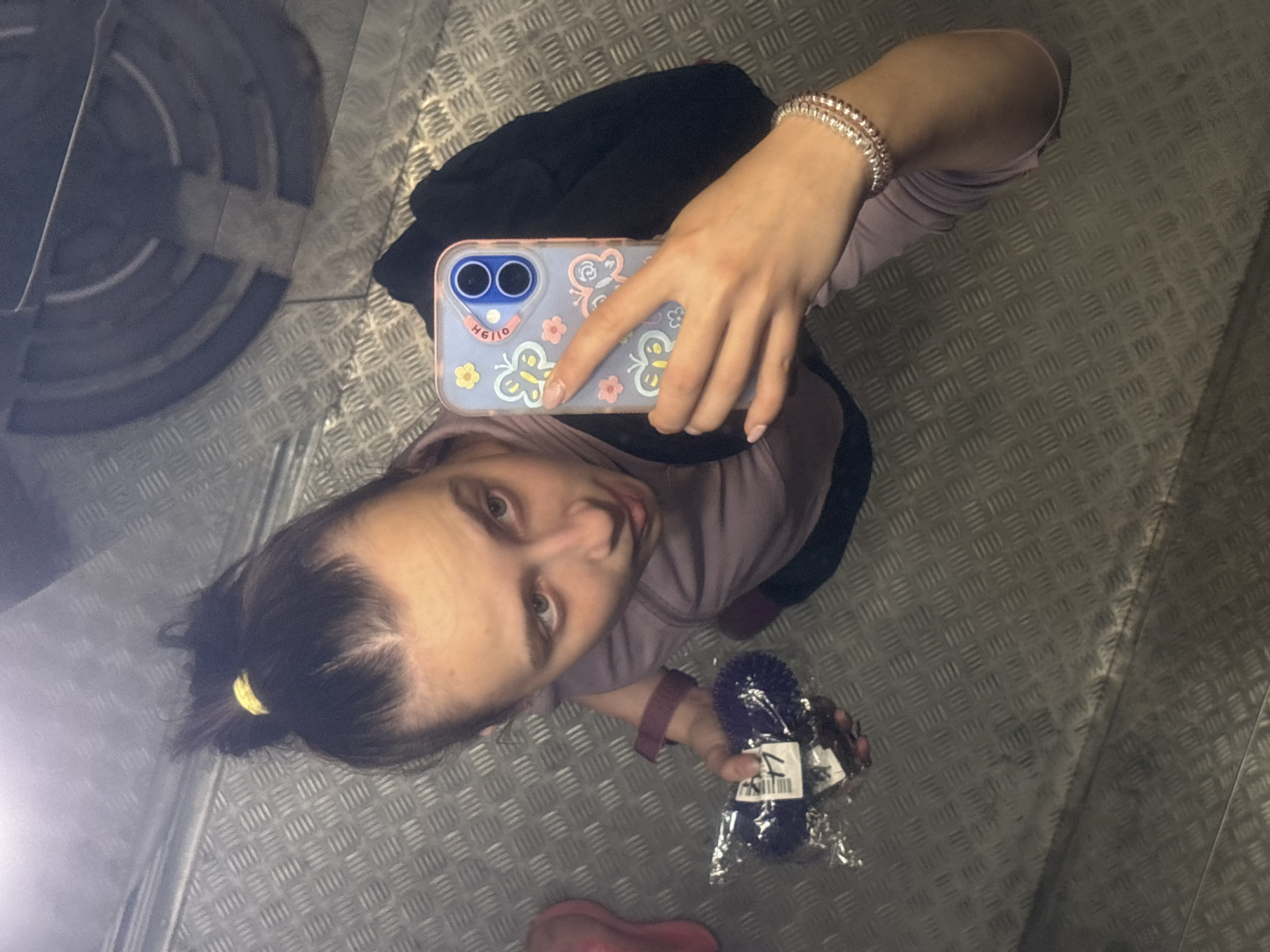 A young woman takes a selfie in a metal-walled elevator. She holds an iPhone with a butterfly case in her right hand, the screen facing away. She wears a lavender long-sleeved shirt and a bracelet on her right wrist. Her hair is pulled back in a ponytail with a yellow scrunchie. In her left hand, she holds a small, dark blue or purple pom-pom with a tag, still wrapped in plastic. The elevator floor is patterned metal. A circular structure (possibly the elevator mechanism) is visible in the upper left corner. A black bag is slung over her shoulder. The location is possibly Perm, Russia.