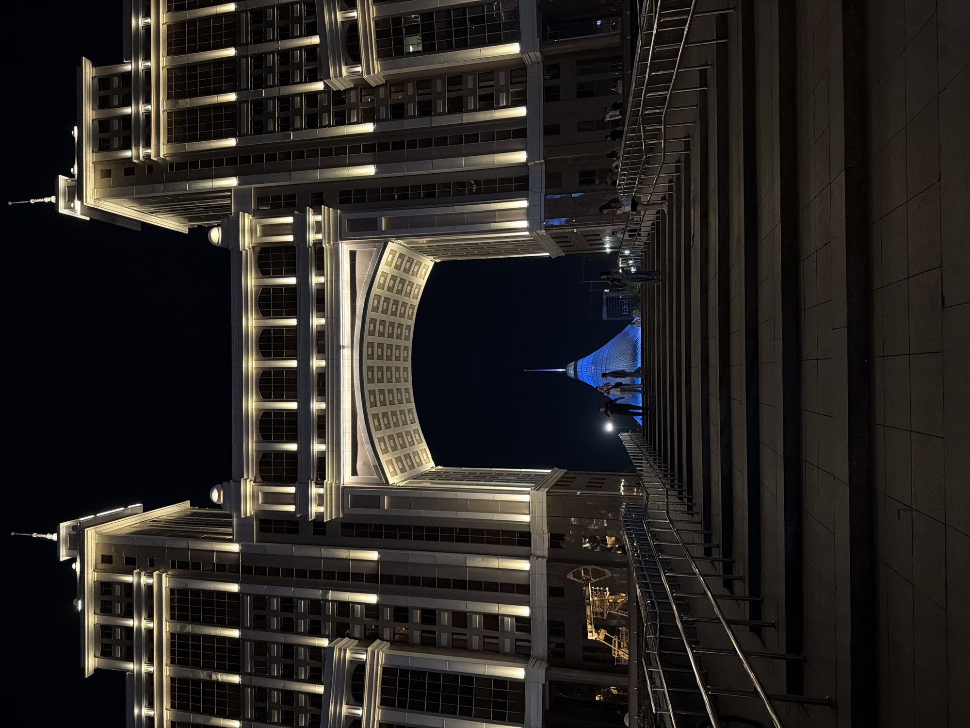 This is a night-time, eye-level, medium shot of the Palace of Peace and Reconciliation in Astana, Kazakhstan, viewed from a grand staircase. The illuminated building, with its distinct pyramid shape topped by a spire, is visible in the background through a large archway formed by two imposing, modern buildings. These flanking structures are tall and rectangular with a classical architectural style, featuring rows of windows and decorative exterior lighting that highlights their verticality and ornamentation. The archway itself is elaborately decorated with a patterned ceiling. On the staircase, several individuals are seen walking up and standing, some appearing to be tourists observing the architecture. The scene is bathed in artificial light from the buildings and street lamps, creating a dramatic contrast with the dark night sky. The staircase is wide and made of dark stone, with metal railings on either side.