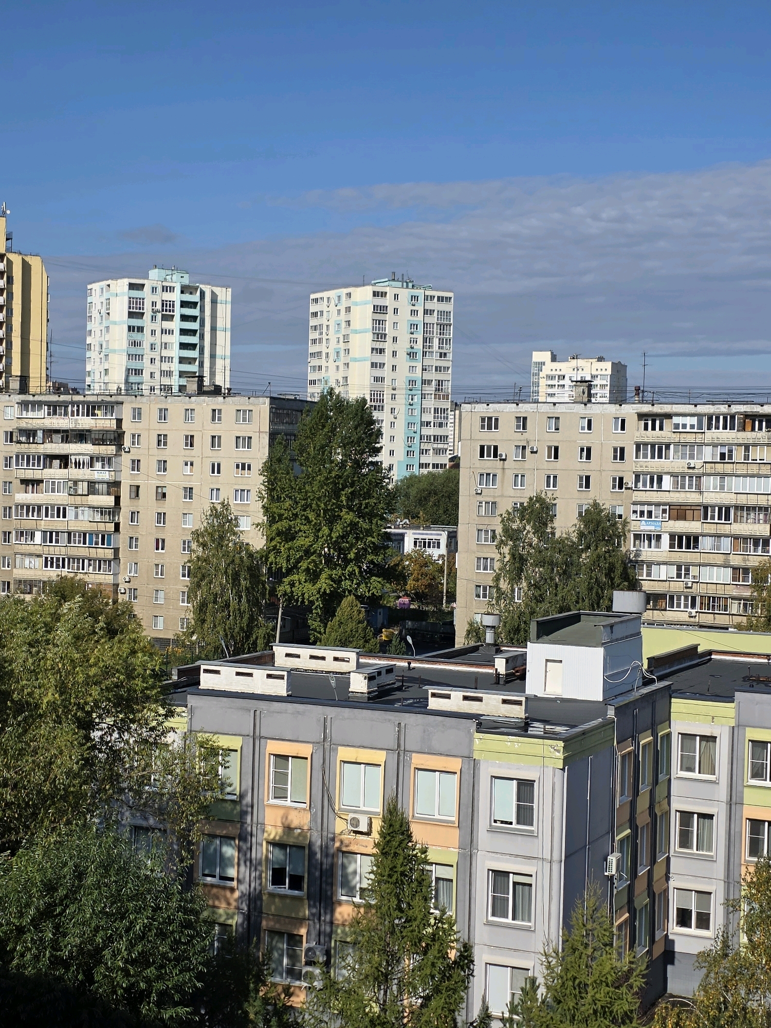 The image depicts a view of residential buildings in Chelyabinsk, Russia, under a partly cloudy sky. The buildings are of varying heights and architectural styles, characteristic of urban development. In the foreground, a lower, multi-story building with a grey facade and colorful accents is visible, partially obscured by lush green trees. Behind this, taller apartment buildings dominate the middle ground. Some of these structures are light-colored with balconies, while others feature more muted tones. Further in the distance, more apartment blocks rise against the sky, some with a pale yellow or light blue trim. The scene suggests a typical urban residential area, likely a district with a mix of older and more recent construction. The foliage indicates it is likely daytime, possibly late summer or early autumn given the green leaves on some trees. There are no people or any discernible activity captured in the frame.
