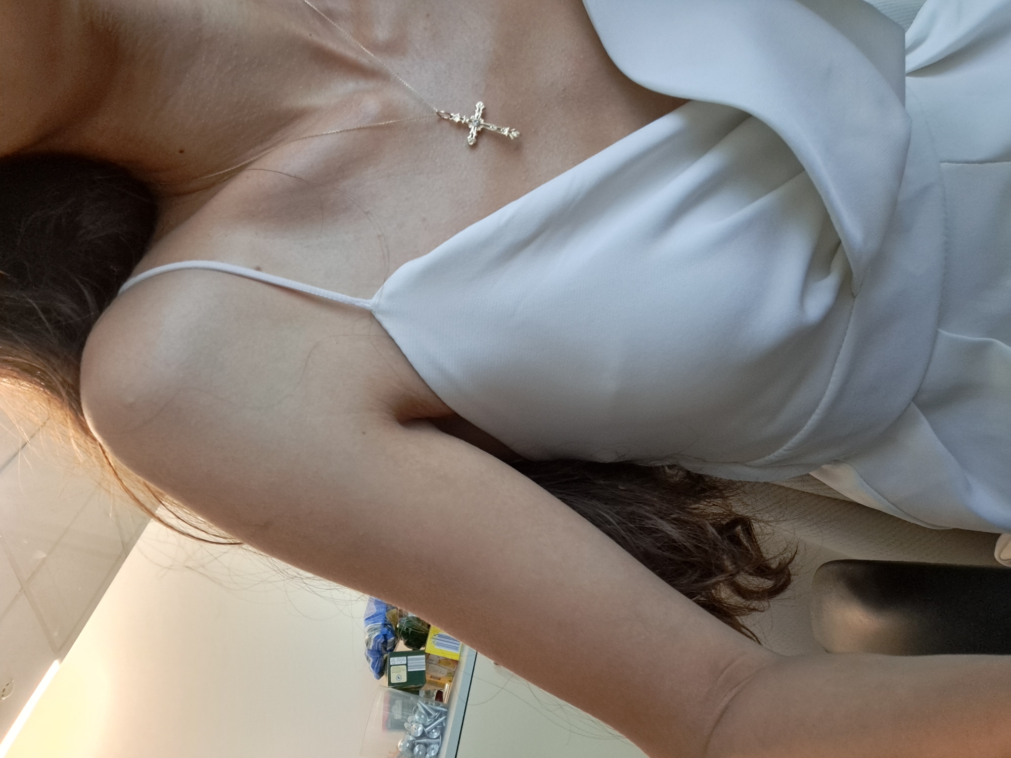 This is a close-up, low-angle shot of a person wearing a white dress with thin spaghetti straps. The person has long, dark brown hair that falls over their shoulder and arm. A delicate silver necklace with a cross pendant is visible around their neck. The lighting suggests an indoor setting, possibly a well-lit room or a studio, with a bright light source coming from the left. The background is blurred, but hints of shelves with various items, possibly packaging or containers, are visible, suggesting a retail or storage environment. The overall impression is intimate and focused on the texture and detail of the dress and jewelry.
