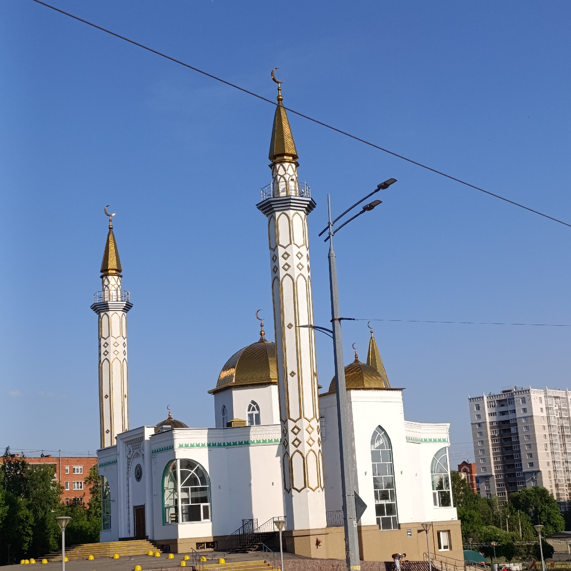 The image features a mosque in Izhevsk, Russia, set against a clear blue sky. The mosque is primarily white with golden domes and accents, featuring two prominent minarets adorned with crescent moons. In the background, there's a mix of urban development, including apartment buildings and trees. In the foreground, are steps leading up to the mosque entrance, streetlights, and overhead wires. A person is visible in the lower right corner.
