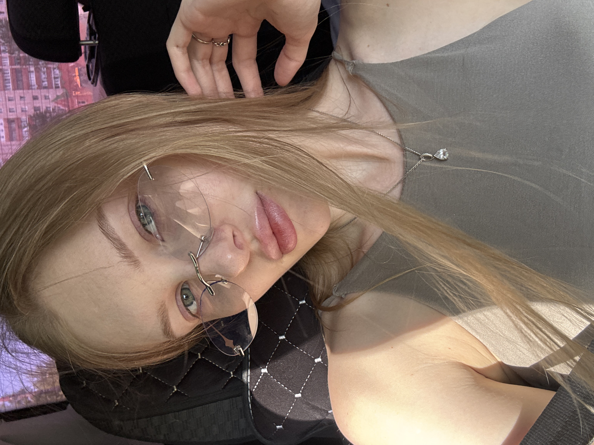 A young woman with light blonde hair and fair skin is the subject of this portrait. She is wearing round, clear-lensed eyeglasses with a unique branch-like design for the temples. A thin silver necklace with a small pendant is visible around her neck. She is wearing a gray top. Her left hand is raised towards her head. She is wearing rings on her fingers. The background is a blurred cityscape with buildings. The image appears to be taken in Perm, Russia. The seating in the picture features a black fabric with a diamond-stitched pattern.