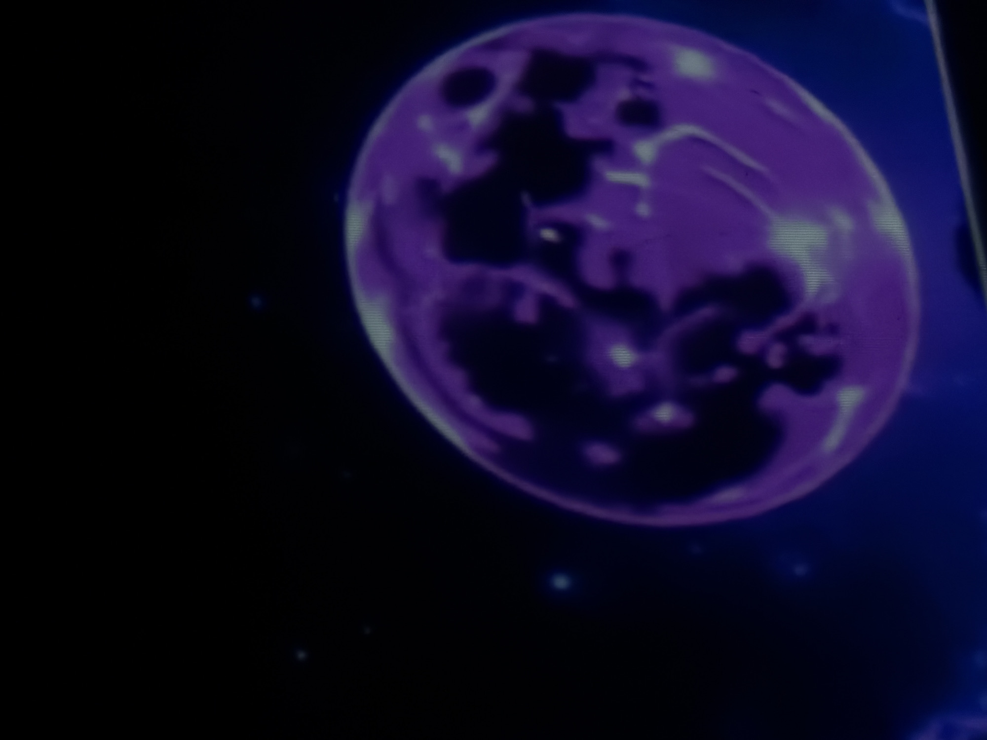 The image is a close-up, slightly blurry photograph of a stylized, luminous purple sphere set against a dark background with faint blue and white speckles, suggestive of a starry night sky or outer space. The sphere has darker, amorphous shapes within it, creating a mottled or marbled appearance, and lighter, glowing areas that suggest internal light or texture. The overall aesthetic is digital and somewhat abstract. There is no identifiable location in the city of Tbilisi, Georgia, or any discernible people, actions, or interactions.