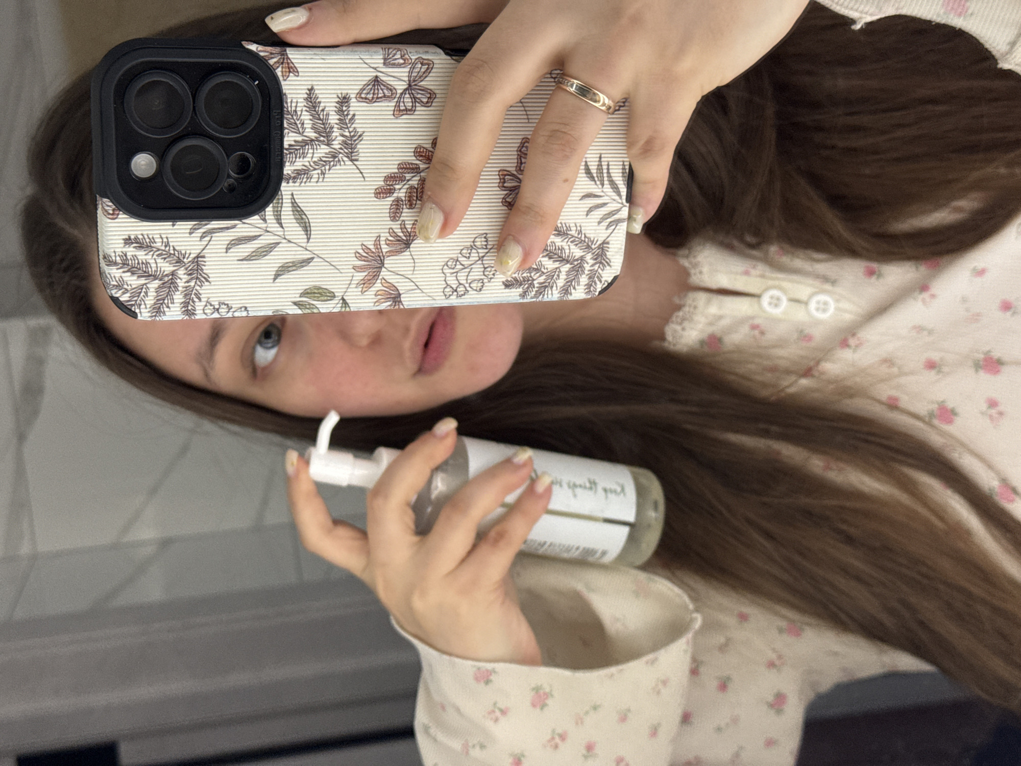 A woman is taking a selfie in an indoor bathroom setting. She is holding a white lotion or soap bottle with a pump dispenser in one hand and a smartphone in the other, obscuring part of her face with the phone. The smartphone has a decorative floral patterned case and a black camera lens housing. She has long brown hair and is wearing a button-down shirt with a small pink floral pattern. The bathroom has light grey tiled walls. There is no indication of Krasnodar, Russia.