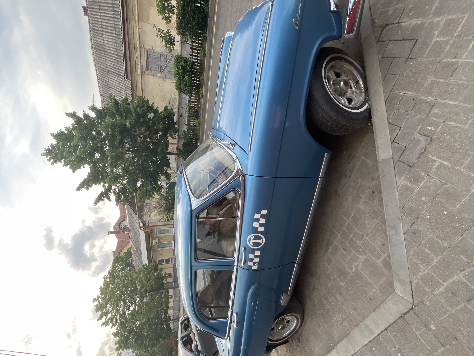 This full-shot photograph captures a light blue vintage GAZ-21 Volga sedan, configured as a taxi, parked on a cobbled surface adjacent to a paved street in Krasnodar, Russia, under an overcast sky.
The central subject is the light blue GAZ-21 Volga sedan, positioned in the lower-middle to right of the frame, angled slightly towards the viewer. The vehicle exhibits a glossy finish and is adorned with extensive chrome trim along its windows, side body, and front fenders. White checkered decals, featuring a white capital 'T' enclosed within a white circle, are prominently displayed on the driver's side rear door, identifying it as a taxi. The car is equipped with multi-spoke chrome or polished alloy wheels, fitted with tires featuring white sidewalls. The front right tire is partially visible and slightly angled. Reflections on the car's windows obscure clear visibility of the interior, but indistinct figures consistent with at least two occupants are discernible. The "Волга" script in Cyrillic is visible on the passenger side front fender.
The background consists of a street scene with several older buildings and mature trees. To the left and behind the taxi, a two-story building with a light-colored, possibly stucco, facade is visible. It features a corrugated metal roof, multiple rectangular windows, and two white external air conditioning units mounted on its exterior. Behind this structure, a building with a reddish-brown tiled roof is partially observed. To the right of the taxi, another weathered, light-colored building with a similar corrugated metal roof features a distinctive, boarded-up wooden shuttered window or door. A low, dark wooden picket fence runs along the front of this building, partially obscured by green bushes and scattered red rose blossoms. Numerous deciduous trees with dense green foliage are distributed throughout the scene, partially obscuring the buildings and the sky. The sky above is bright but covered by diffuse grey and white clouds. A dark-colored modern car is partially visible parked behind the blue taxi on the far left. The paved street runs parallel to the cobbled parking area, separated by a concrete curb.