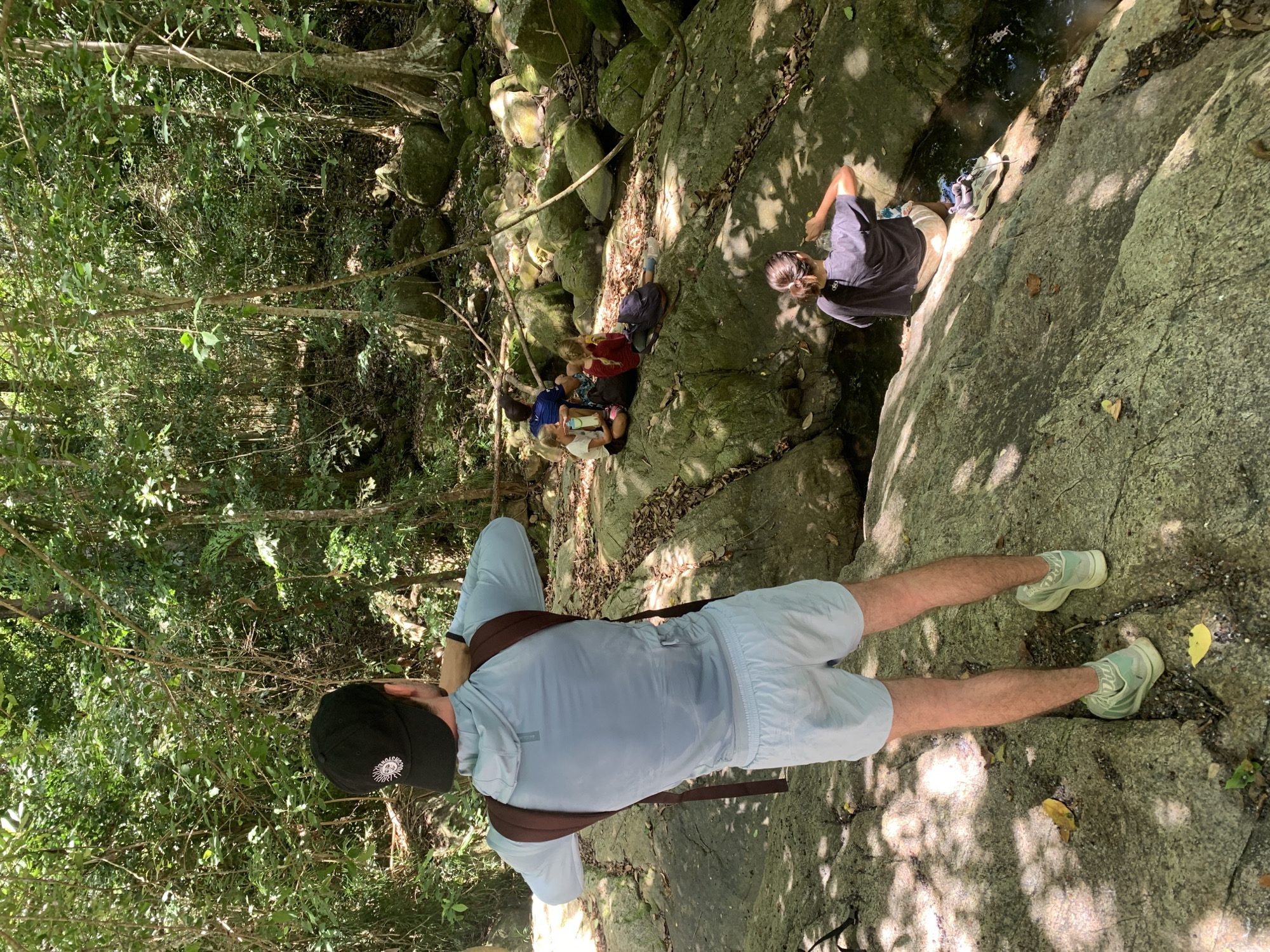 The image shows a group of people in a natural, rocky environment that may be a park or hiking area near Baan Lipa Noi, Thailand. A man stands in the foreground on a large rock, facing a group of people seated near a small stream or pool of water. He wears a light blue shirt, shorts, a black cap, a backpack, and light green sneakers. The group by the water consists of several individuals, some sitting and some standing. The environment is lush with greenery and rocks, with sunlight filtering through the trees. The scene suggests a casual outing or hike with people taking a break near the water.