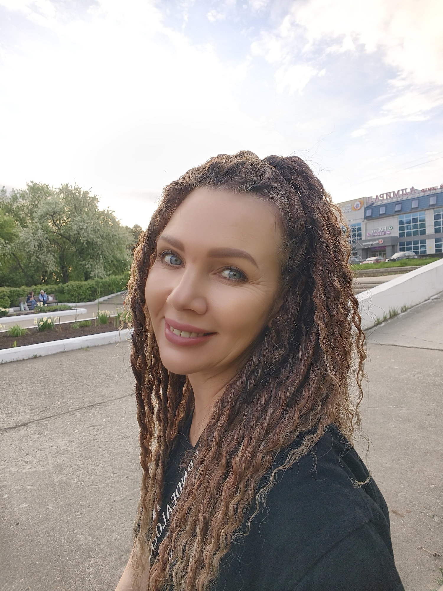 A woman with brown and blonde dreadlocks is smiling and looking directly at the camera. She is wearing a black t-shirt with white lettering. The background consists of a paved area, a grassy area with trees and flowers, and a building with a sign that reads "ЛАМПИЛА" in Cyrillic characters. In the grassy area, two people are sitting on a bench. The location appears to be a park or outdoor area in a city.