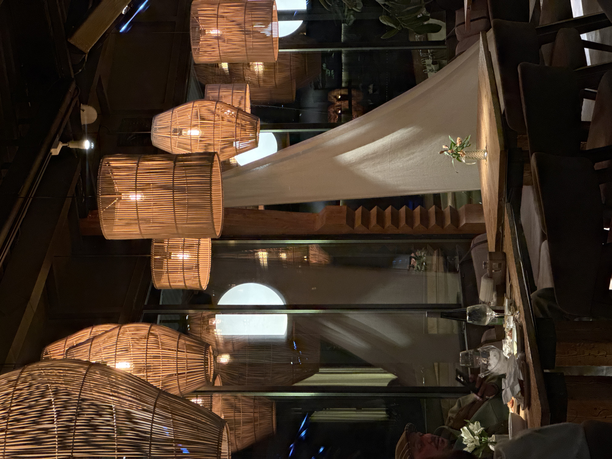 The image captures an intimate indoor dining or lounge area, characterized by warm, dim lighting and a blend of natural and modern aesthetics. Several cylindrical and elongated pendant lamps, crafted from woven bamboo or wicker, hang from a dark ceiling, casting a soft, golden glow throughout the space.
A long, draped white fabric element extends diagonally from the upper left to the lower right of the image, serving as a subtle architectural accent. Below this, large glass partitions or windows reflect the interior lighting and elements, including multiple instances of the radiant lamps and parts of the ceiling structure. Through the glass, shadowy reflections of the venue's interior or ambient light sources are subtly visible.
On the right side of the frame, dark upholstered chairs are arranged, suggesting dining tables or seating areas. A wooden surface, possibly a table or bar top, is visible, adorned with a small vase holding a few delicate flowers. Further down, a dining setup includes wine glasses, a wine bottle, and crumpled napkins, hinting at a meal or social gathering. A person wearing a hat is partially visible and out of focus in the lower right corner, suggesting a patron enjoying the setting.
The overall ambiance is cozy and sophisticated, indicative of an evening setting. The decor combines rustic charm from the natural lamp materials and wooden elements with a contemporary feel from the glass and sleek furniture. No text is visible in the image.