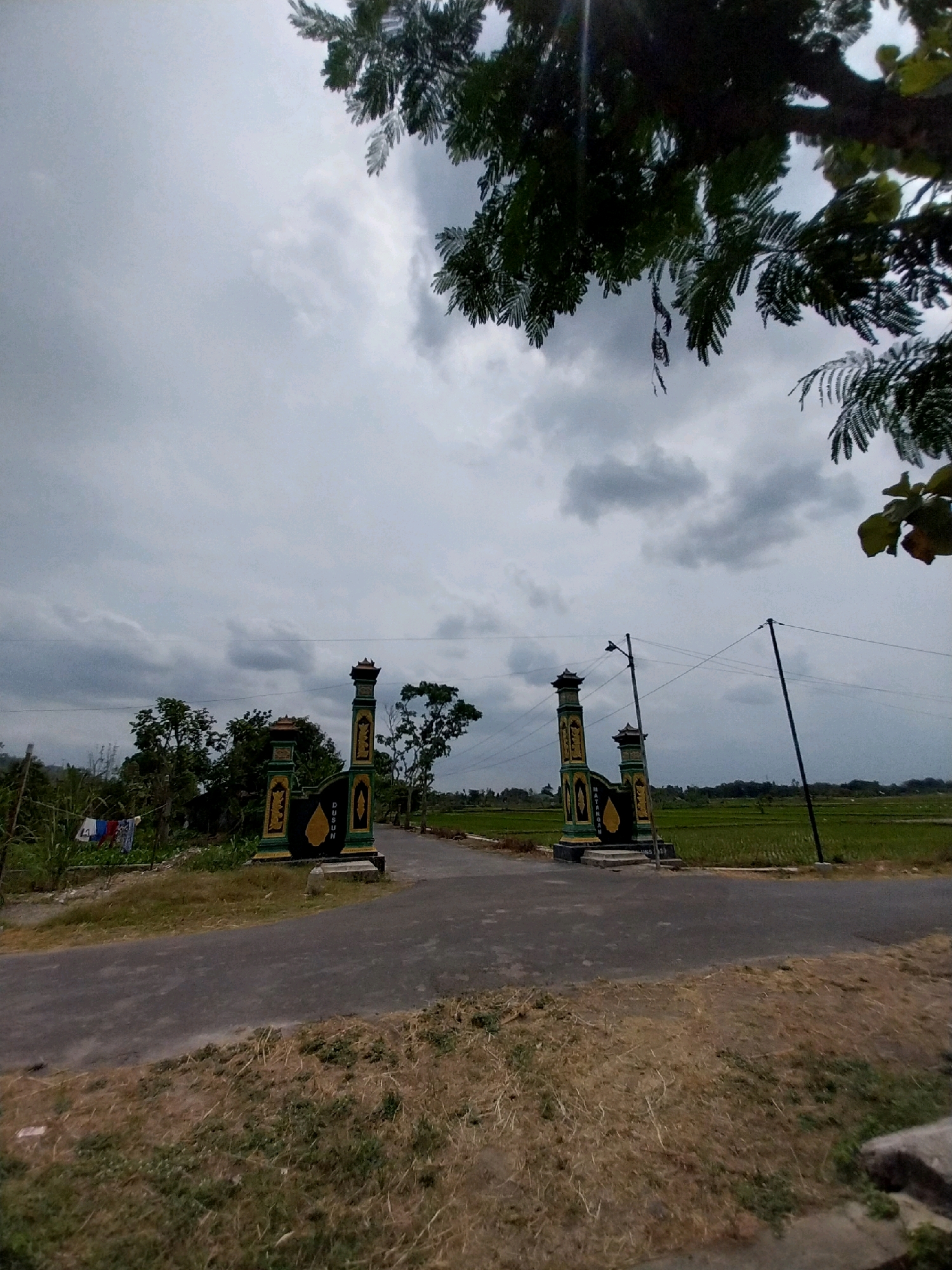 The image depicts a rural scene in Cucukan, Indonesia, under a cloudy sky. In the foreground, a paved road runs towards an ornate gateway, flanked by two pillars adorned with green, yellow, and black designs. The gateway bears inscriptions in Indonesian, indicating "DUSUN" and "MATTANAN". Beyond the gateway, a vibrant green rice field stretches towards the horizon, with scattered trees and vegetation. A utility pole with electrical wires stands to the right of the gateway. In the background, a cluster of trees and a small structure are visible on the left side of the road, with laundry hanging on a line. The overall atmosphere is overcast, suggesting a potentially humid climate.