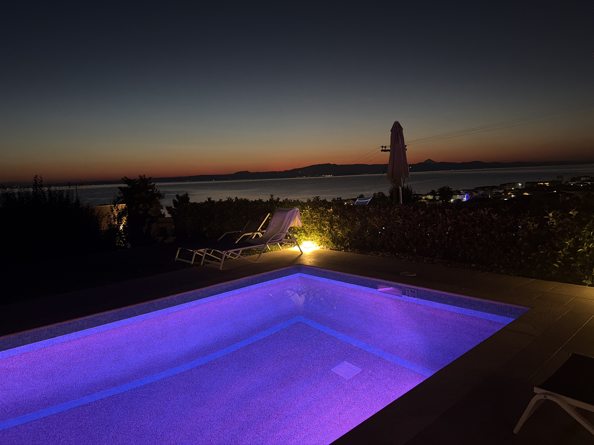 The image captures a twilight scene at a swimming pool, likely in Pallini Municipal Unit, Greece. The sky transitions from a deep blue overhead to hues of orange and red near the horizon, indicating the sun has recently set. A calm body of water, possibly the Aegean Sea, stretches across the background, bordered by distant hills and a town with illuminated buildings. In the foreground, a swimming pool glows with vibrant purple lighting, its tiled bottom visible beneath the illuminated water. Two lounge chairs, one with a white cushion, are positioned on the deck beside the pool. A closed patio umbrella stands nearby, partially obscured by lush green foliage that lines the edge of the property. The overall atmosphere is serene and tranquil, characteristic of a vacation setting at dusk.