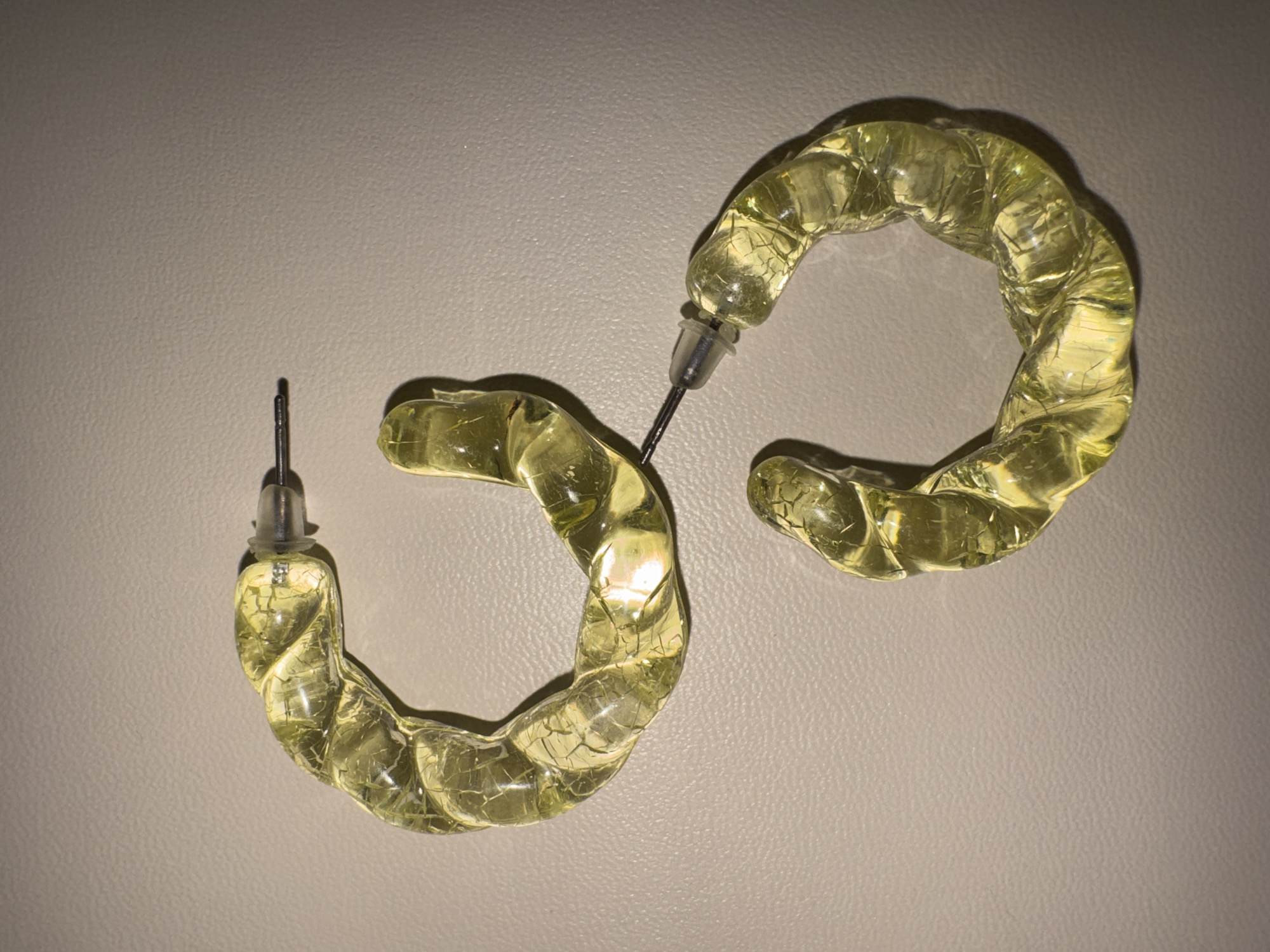 The image shows a pair of yellow, translucent, hoop earrings with a twisted design. The earrings are made of a material that appears to be acrylic or resin, with visible internal crackling or inclusions. Each earring has a silver post and a clear plastic backing. One earring is positioned to the left of the frame, with its post pointing upwards and slightly to the left. The other earring is to the right and slightly above the first, with its post pointing downwards and slightly to the right. The earrings are lying on a textured, light-colored surface that resembles leather or a similar material. The lighting in the image creates subtle shadows and highlights on the earrings, emphasizing their translucent quality and twisted form. There is no text visible in the image. The setting is a flat surface, likely a tabletop or display area. The image is a close-up, top-down view of the jewelry.