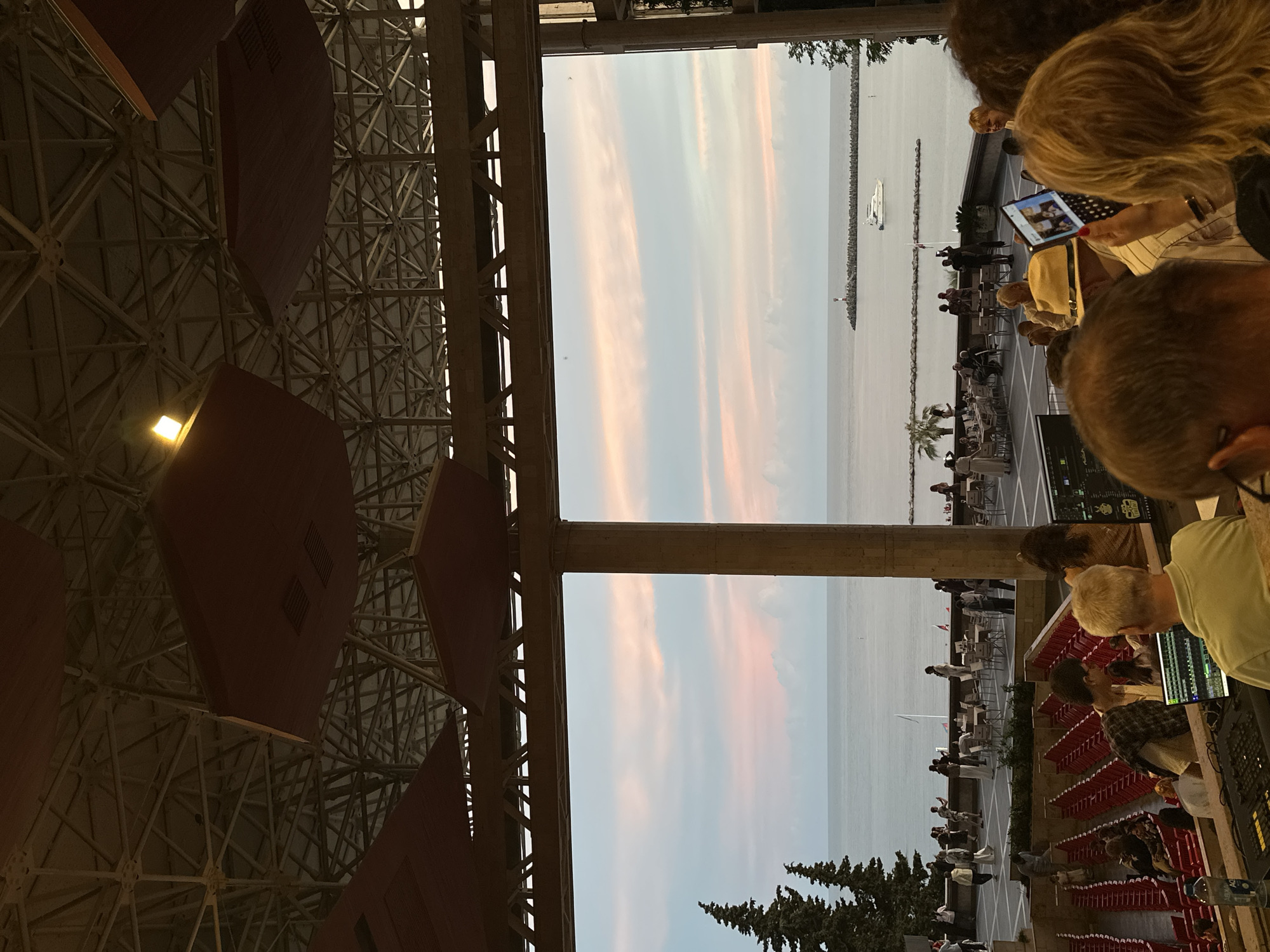 This image captures an outdoor amphitheater in Sochi, Russia, with the Black Sea in the background at dusk. The sky displays a soft gradient of pinks and blues, indicating sunset. In the foreground, a person is operating a soundboard, facing away from the camera. Rows of empty red seats fill the lower part of the amphitheater.
Further back, on a paved terrace overlooking the sea, several people are seated at tables, some appearing to be dining or socializing. Trees, including tall conifers, dot the landscape to the left. A lighthouse and a white yacht are visible on the water, with a breakwater extending into the sea. The amphitheater's roof structure, a complex metal framework with suspended reddish panels, is prominent at the top of the image. The overall scene suggests an evening event or gathering, possibly a concert or performance, taking place in a scenic coastal location.