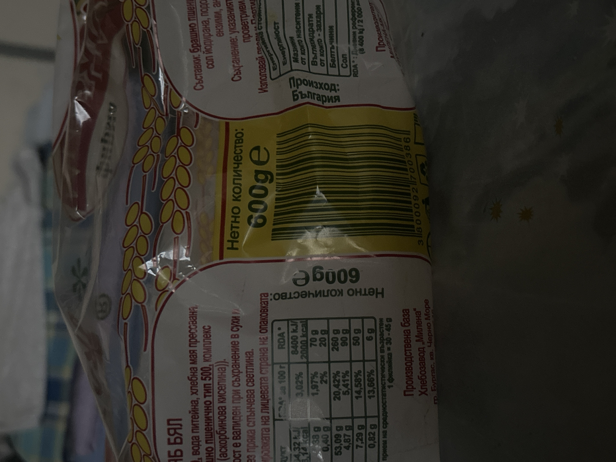 The image displays a translucent plastic package of a food product, likely bread or a similar baked good, with printed labels predominantly in Bulgarian. The package is slightly crumpled, suggesting it might be in a casual setting like a kitchen or store. Stylized wheat grains are part of the packaging design.
Key information visible on the package includes:
* **Product Name:** "АБ БЯЛ" (AB BIAL).
* **Net Quantity:** "Нетно количество: 600ge" (Net quantity: 600g).
* **Ingredients (Съставки):** Wheat flour (тип 500), drinking water, pressed baker's yeast, iodized salt, and an enzyme complex (ascorbic acid).
* **Storage (Съхранение):** Instructions advise storing in a dry and ventilated place, away from direct sunlight.
* **Origin (Произход):** Bulgaria.
* **Production Base (Производствена база):** "Хлебозавод „Милена" гр. Бургас, кв. Черно Море" (Bakery "Milena", Burgas, Black Sea region).
A nutritional information table provides values per 100g and as a percentage of the Recommended Daily Allowance (RDA), which is based on 8400 kJ / 2000 kcal. Categories listed include Energy, Fats (with saturated fats specified), Carbohydrates (with sugars specified), Protein, and Salt. For example, 100g contains 53.09g of carbohydrates and 7.29g of protein. A serving size is indicated as "1 филийка = 30-45 g" (1 slice = 30-45 g). A barcode is also visible on the packaging. The background is indistinct and appears dimly lit, indicating an indoor environment.