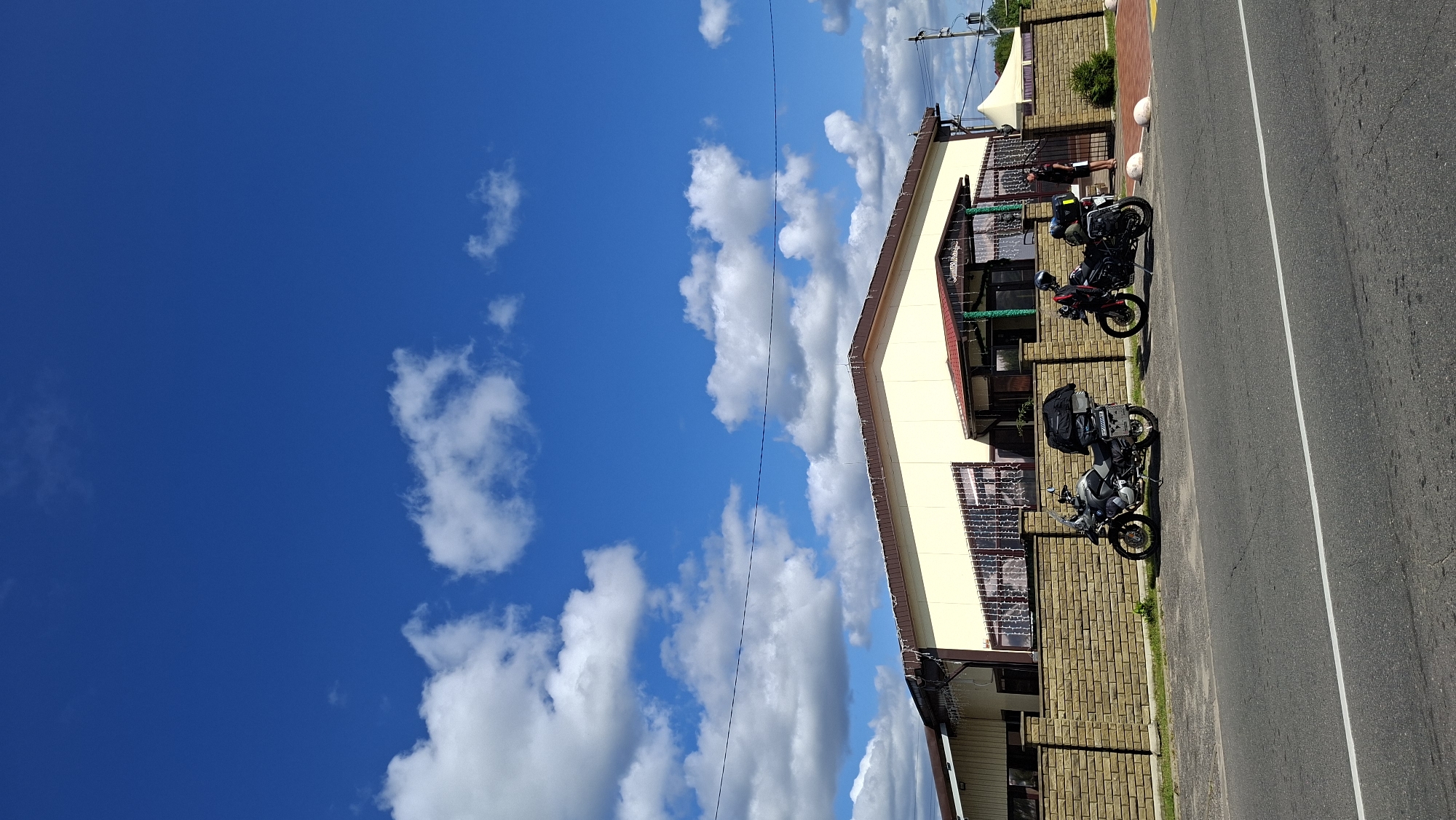 In Myadzyel, Belarus, a light-yellow building with a brown roof stands behind a brick wall. Two motorcycles are parked on the paved area in front of the wall. The motorcycle on the left is silver and black, equipped with luggage racks. The motorcycle on the right is black and red, also with luggage racks. A person in a red jacket is standing next to the right motorcycle, facing away from the camera. Another person in a black shirt and white shorts stands further right, near a white tent. The scene is set against a backdrop of a bright blue sky with scattered white clouds. A paved road runs in front of the motorcycles.