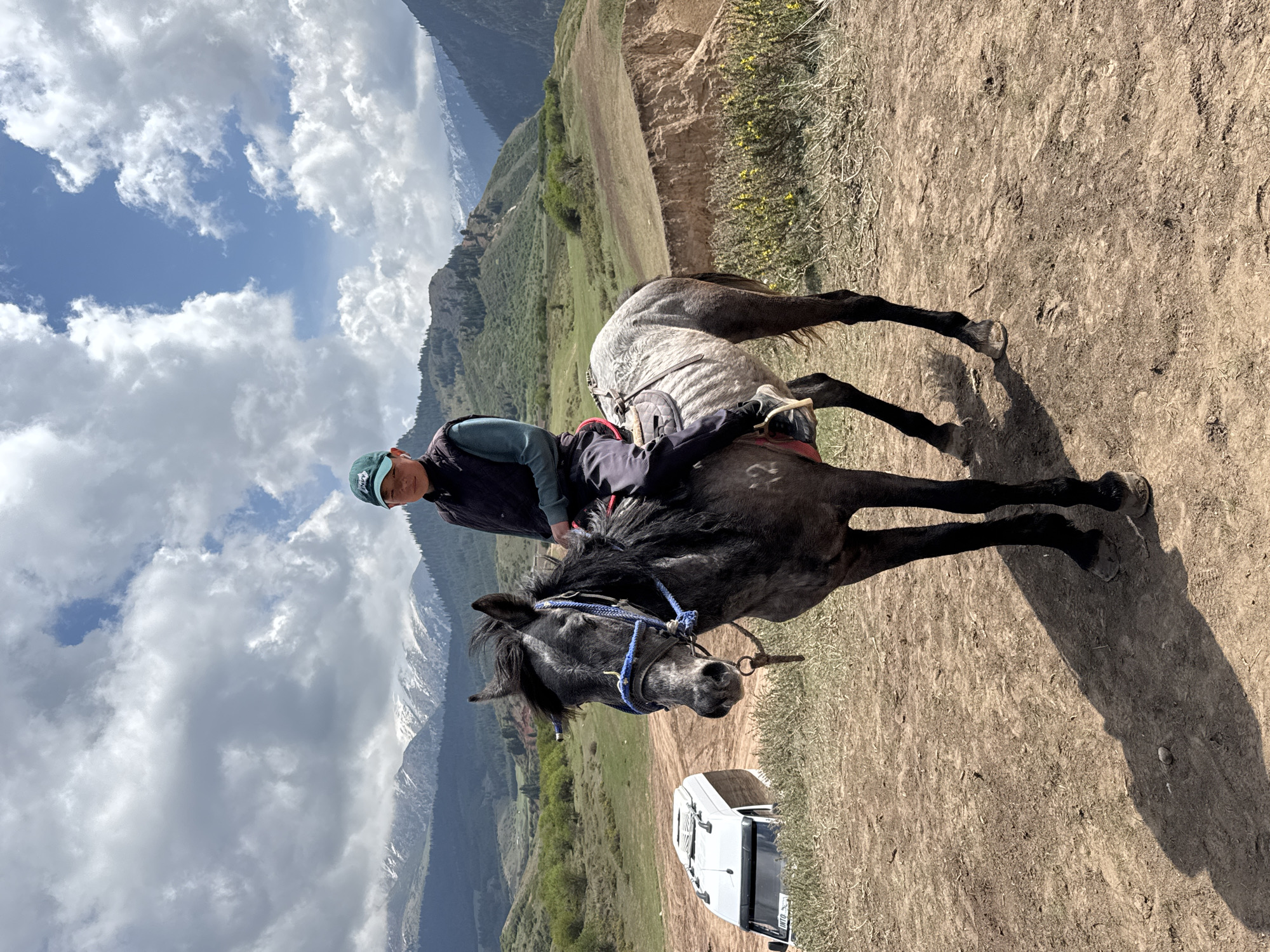 A person is depicted riding a dark grey or black horse in a scenic mountainous landscape in Jeti-Ögüz, Kyrgyzstan. The image is brightly lit, with strong shadows indicating midday or early afternoon.
The central subject is an individual wearing a blue cap, a dark vest, and a light-colored shirt, seated on the horse. The horse is fitted with a saddle and a blue bridle and reins, with its head turned slightly towards the viewer. The rider appears to be resting or paused on a dirt path.
In the background, majestic snow-capped mountains rise under a partly cloudy sky. The lower slopes of the mountains are covered with green vegetation. The midground consists of rolling hills with brown and sparse green growth. A patch of yellow flowers is visible on a bank to the right. The sky above is a clear blue with scattered white cumulus clouds.
Partially visible behind the horse, to the lower right, is a white vehicle, possibly an SUV or camper van, with the word "INTO" visible on its side. The scene conveys a sense of outdoor adventure amidst rugged natural beauty.