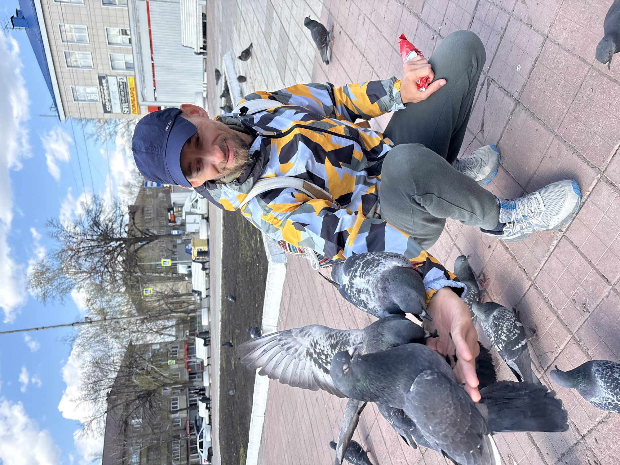 The media file captures a cheerful scene of a man feeding pigeons in what appears to be a city square or public area in Ulyanovsk, Russia. The man is squatting on a reddish-brown tiled pavement, smiling towards the viewer. He wears a dark blue baseball cap with "FOX LTL" visible, a distinctive windbreaker jacket featuring a geometric pattern in yellow, light blue, and black, dark green pants, and light-colored athletic shoes, one displaying the "ASICS" brand. In his left hand, he holds a red packet, possibly containing food for the birds.
Several pigeons surround him, with multiple birds actively feeding directly from his outstretched right hand and perched on his arm and shoulder. Other pigeons are on the ground nearby. The background features a multi-story building with numerous windows and several commercial signs, some in Cyrillic, including "COMPONENTS," "SAPPHIR SHOP," and "ПОМБАРД." Bare trees, suggesting late winter or early spring, line the street. The sky is bright blue with scattered white clouds, indicating a clear, sunny daytime. The man's smile suggests enjoyment of the interaction with the birds.