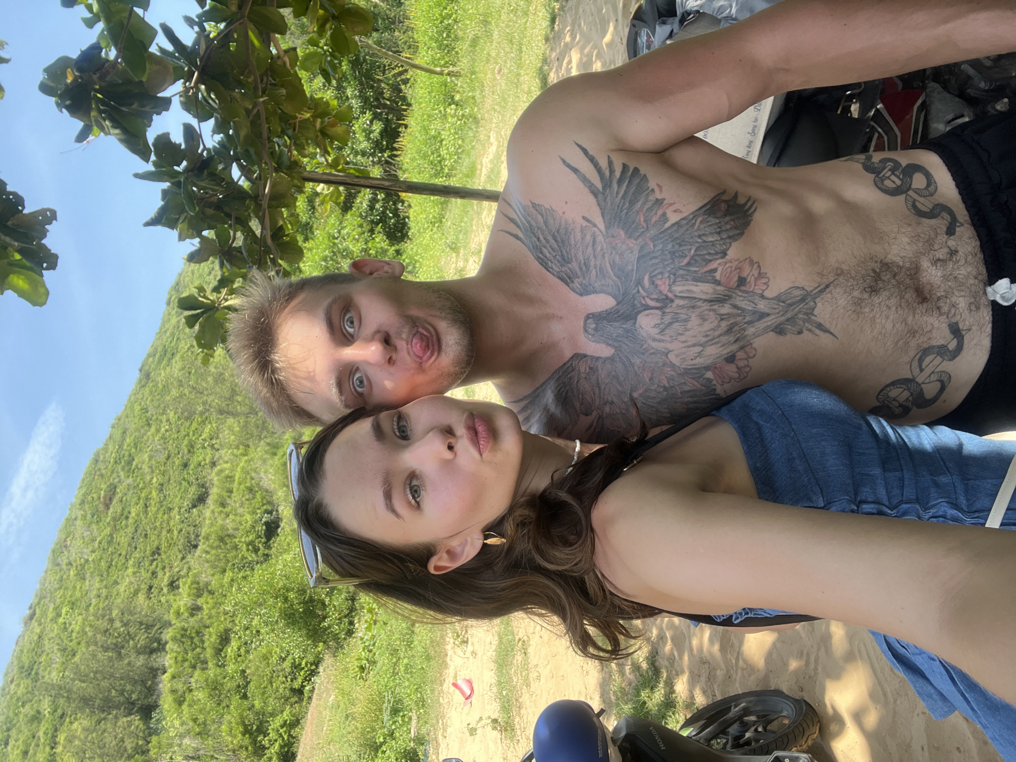A selfie photograph features a fair-skinned woman with long brown hair and a fair-skinned man with short blond hair. The man has a large tattoo of a bird on his chest and abdomen, as well as a snake tattoo. He is shirtless and making a face with his tongue out. The woman is wearing a blue top. They are positioned in front of a lush green hillside, and a tree branch is visible in the upper left corner. A motorcycle is partially visible in the foreground. The photo appears to have been taken outdoors in daylight, possibly on the side of a road. The location is Hòa Xuân, Vietnam.