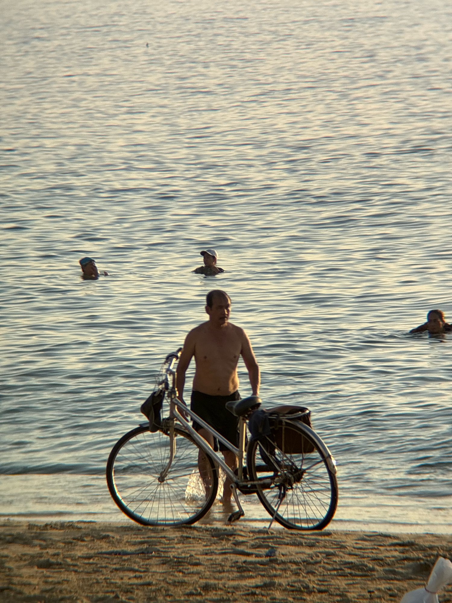 A man with a bare torso and black shorts stands on the beach beside a silver bicycle with a brown saddlebag. He is looking towards the water. In the water, there are at least four other people visible, some with their heads above the surface, and one appears to be swimming. The water is calm with gentle ripples, reflecting the light of what appears to be late afternoon or early morning sun. The beach in the foreground is sandy and appears somewhat dry. The scene suggests a recreational setting, likely a public beach in Nha Trang, Vietnam, where locals and visitors engage in water activities and enjoy the coastline.