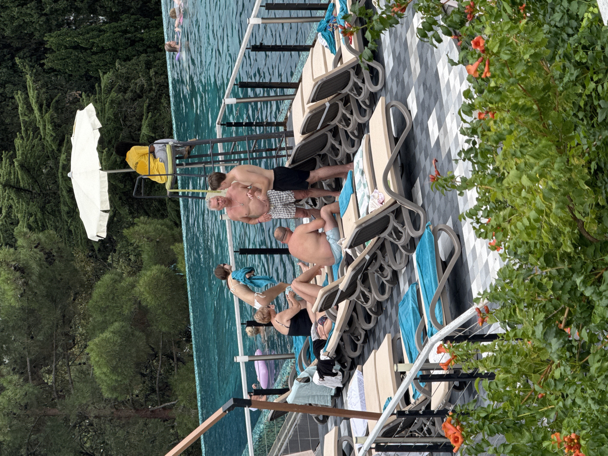 This image depicts a scene at a swimming pool area, likely an outdoor resort or hotel, with lush green trees in the background and flowering orange plants in the foreground. In the mid-ground, several people are present. A lifeguard in a yellow shirt sits on a tall chair under a white umbrella, overlooking the pool. Several individuals are in the pool, with at least two on inflatable rings. On the poolside deck, there are numerous lounge chairs, some occupied by people. One group of individuals appears to be in conversation near the pool's edge. The overall atmosphere suggests a relaxed recreational setting. The location is identified as Yalta, Ukraine, implying this is a scene from a Ukrainian resort.