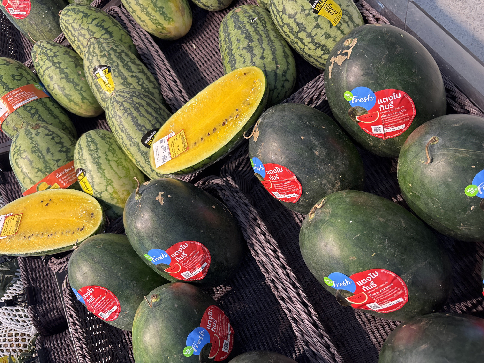 The image shows a collection of watermelons in a gray woven basket, likely at a market or grocery store in บ้านบางรักษ์, Thailand. There are two types of watermelons: oblong, light green watermelons with darker green stripes and darker green, rounder watermelons. Some of the oblong melons have yellow stickers on them, and one is cut open to reveal yellow flesh. The rounder, darker green watermelons are labeled with a red sticker that says "KINNAREE WATERMELON" in Thai and English, along with the claim that it has red flesh and is sweet. The stickers also indicate that the melons are a product of Sakon Nakhon province. One of the rounder melons is cut open and reveals yellow flesh.