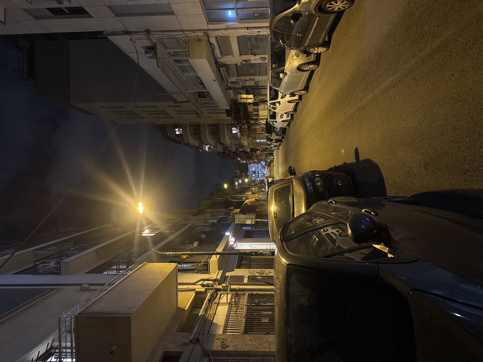 The image depicts a narrow street at night in Tel Aviv, Israel. Cars are parked tightly along both sides of the street. The buildings on either side are multi-story residential apartments, with balconies and window air conditioning units visible. A street lamp illuminates the scene, casting a bright light down the road. The sky is dark, suggesting it is late at night. There is no evidence of people. The perspective is looking down the street, with a slight tilt.