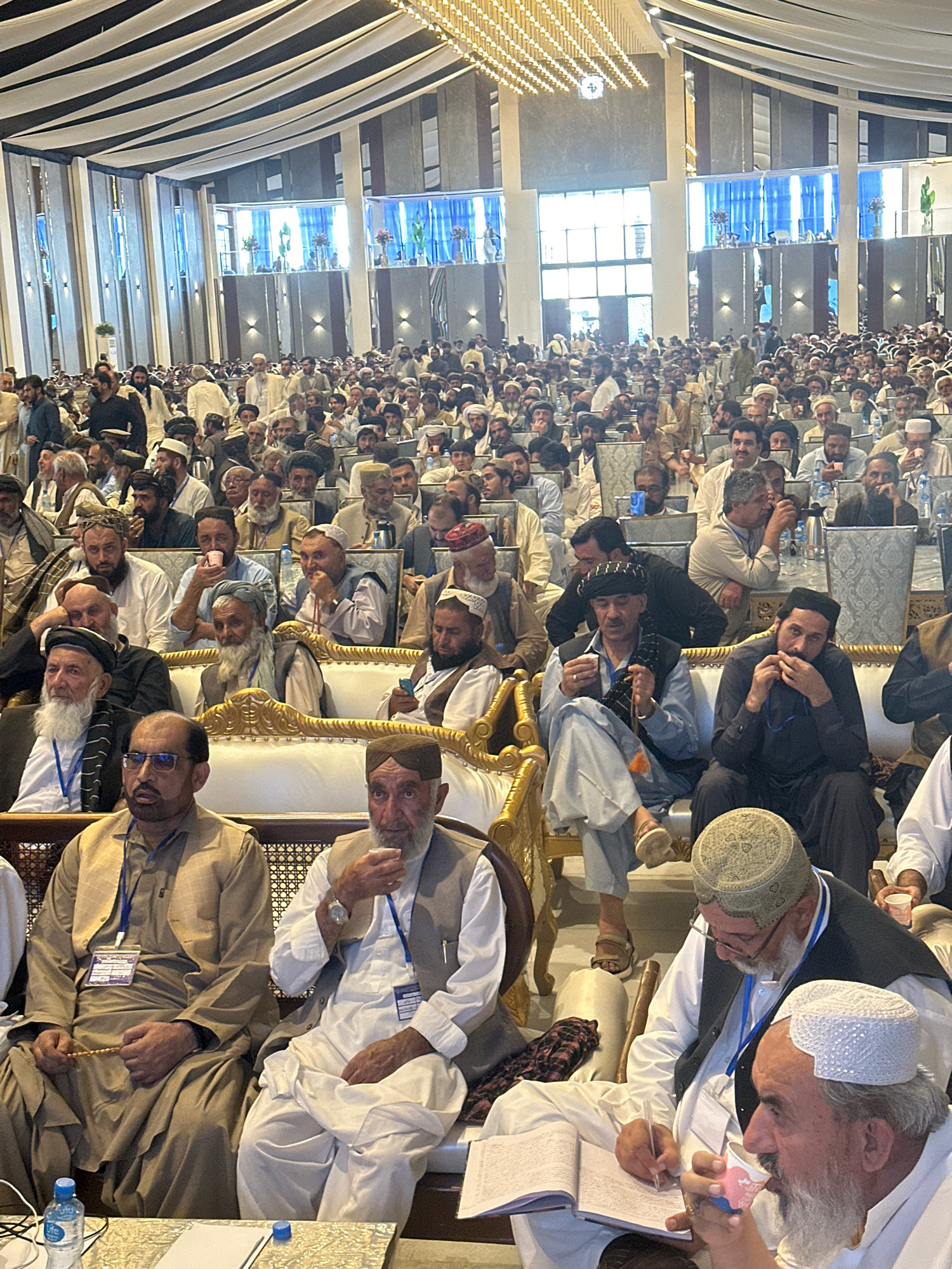 The image depicts a large gathering of men in a spacious indoor venue, likely a conference hall or event space, with a prominent, elaborately decorated ceiling featuring intricate lighting. The men are predominantly dressed in traditional Islamic attire, including turbans, caps, and long robes, with many wearing lanyards and badges, suggesting an organized event.
The seating arrangement consists of rows of chairs and some ornate, cushioned benches, indicating a formal setting. Several individuals are seated in the foreground, engaged in various activities such as holding prayer beads, drinking from cups, writing in notebooks, and conversing. The overall atmosphere appears to be one of a serious assembly, possibly a religious or political conference.
The location is identified as Quetta City Tehsil, Pakistan. This context suggests the event is taking place within the cultural and socio-religious landscape of Balochistan province. The presence of numerous attendees, the formal setup, and the attire of the participants are consistent with such gatherings in the region. The lighting in the venue is bright, illuminating the space and casting shadows, with a mix of natural light from windows and artificial lighting from the overhead fixtures.