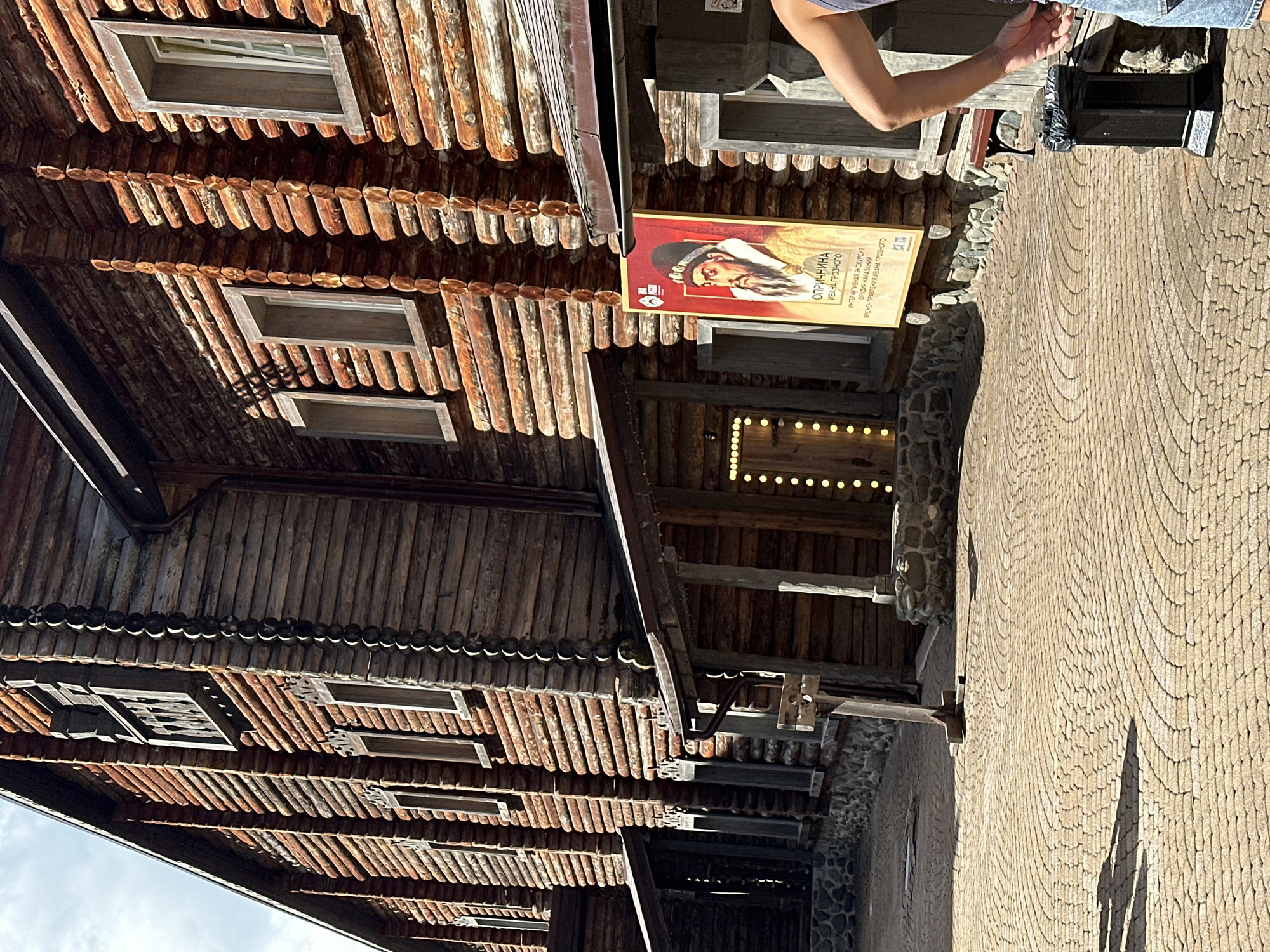 The image shows a wooden building with log construction and several windows. In front of the building, there is a cobblestone pavement with a circular pattern. A poster depicting a man in historical attire is displayed on the wall of the building. There is also a trash can and a person's arm and shoulder visible on the right side of the image. The scene appears to be outdoors on a sunny day. The building resembles traditional Russian architecture, possibly part of an ethnographic park or museum. The poster's text, "ОПРИЧНИНА ИВАНА ГРОЗНОГО" (Oprichnina of Ivan the Terrible), suggests a historical exhibition or attraction. This location could be in the city of Estosadok, Russia, which is known for its natural beauty and cultural attractions.