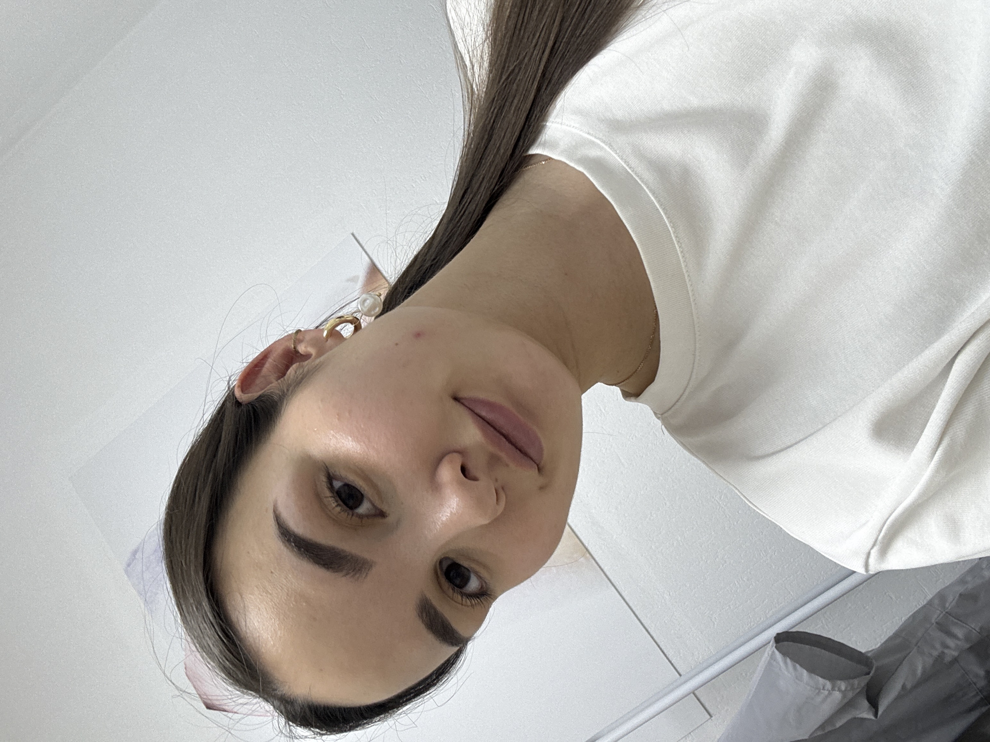 A low-angle close-up shot of a woman with dark hair, fair skin, and brown eyes. She is wearing a white t-shirt. She has a gold earring with a pearl in her left ear. The background consists of a white wall and what appears to be a partially visible poster. The location is Chaikovsky, Russia.