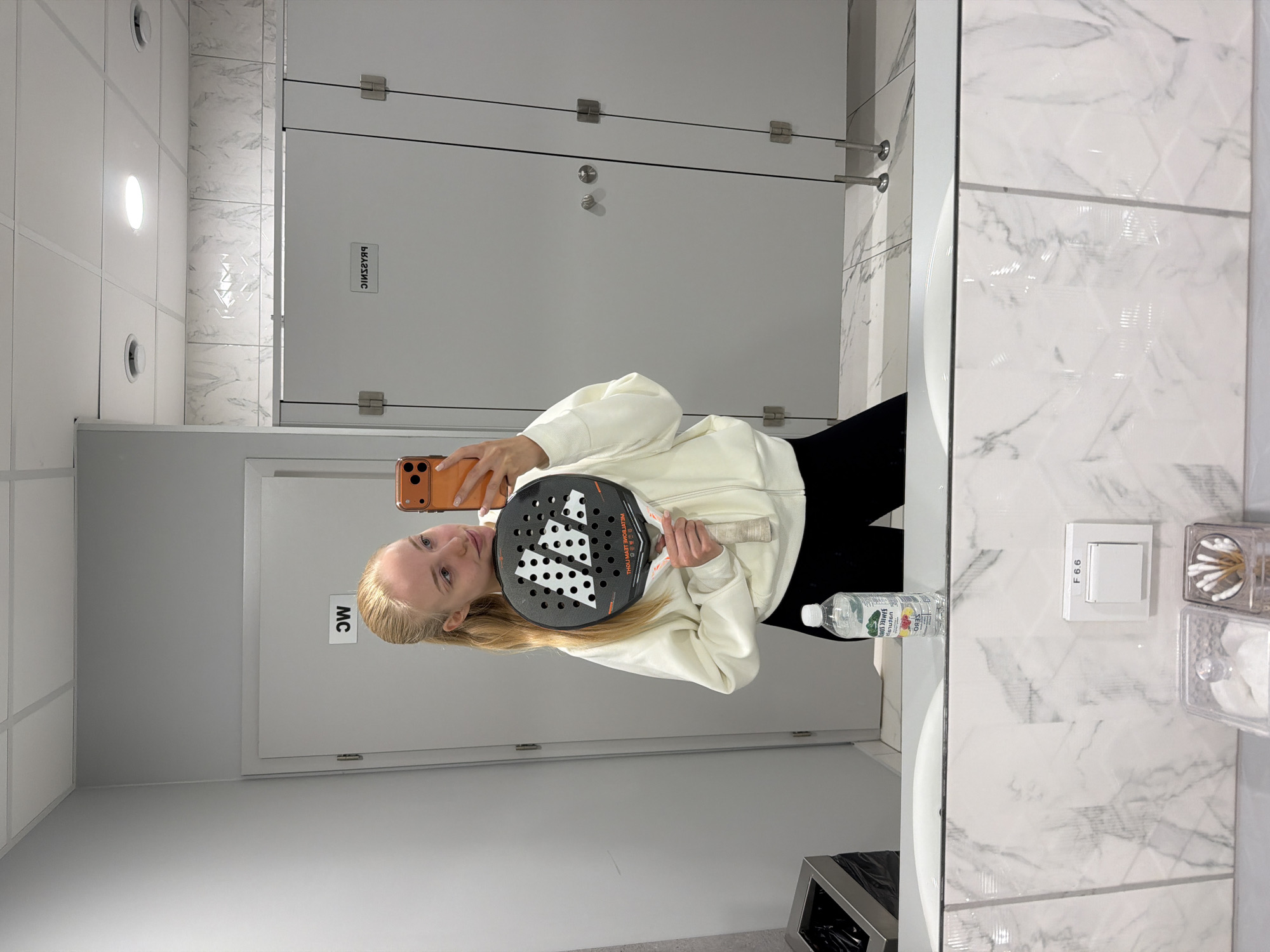 A young woman is taking a selfie in a public restroom. She is holding a padel racket and a phone. She is wearing a white sweatshirt and black pants. There is a bottle of water on the counter. The restroom has white and gray tiled walls, white doors, and a stainless steel trash can.