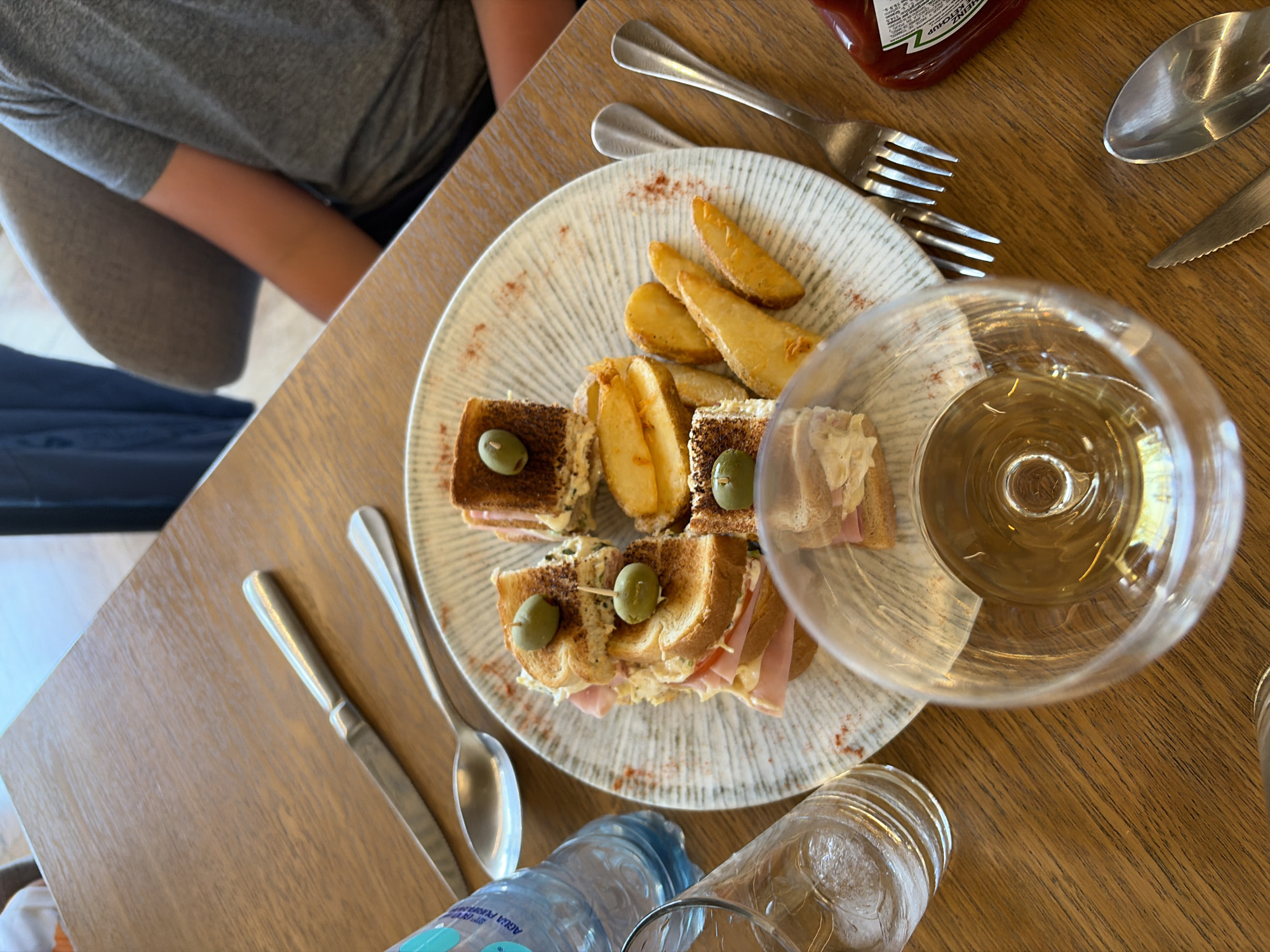 A high-angle shot displays a partially occupied wooden table, likely indoors. The table is set with a plate containing tapas-style sandwiches, each garnished with an olive, and accompanied by potato wedges. Adjacent to the plate is a glass of white wine, a water bottle, and an empty glass. Eating utensils (fork, knife, spoon) are positioned around the plate. A bottle of Heinz ketchup is visible towards the upper right. The presence of tapas suggests a Spanish-influenced cuisine. Given the prompt's mention of Puebla, Mexico, it is reasonable to infer this is a restaurant or cafe in Puebla. One person is partially visible, wearing a gray shirt and seated in a gray chair. No interaction is discernible.