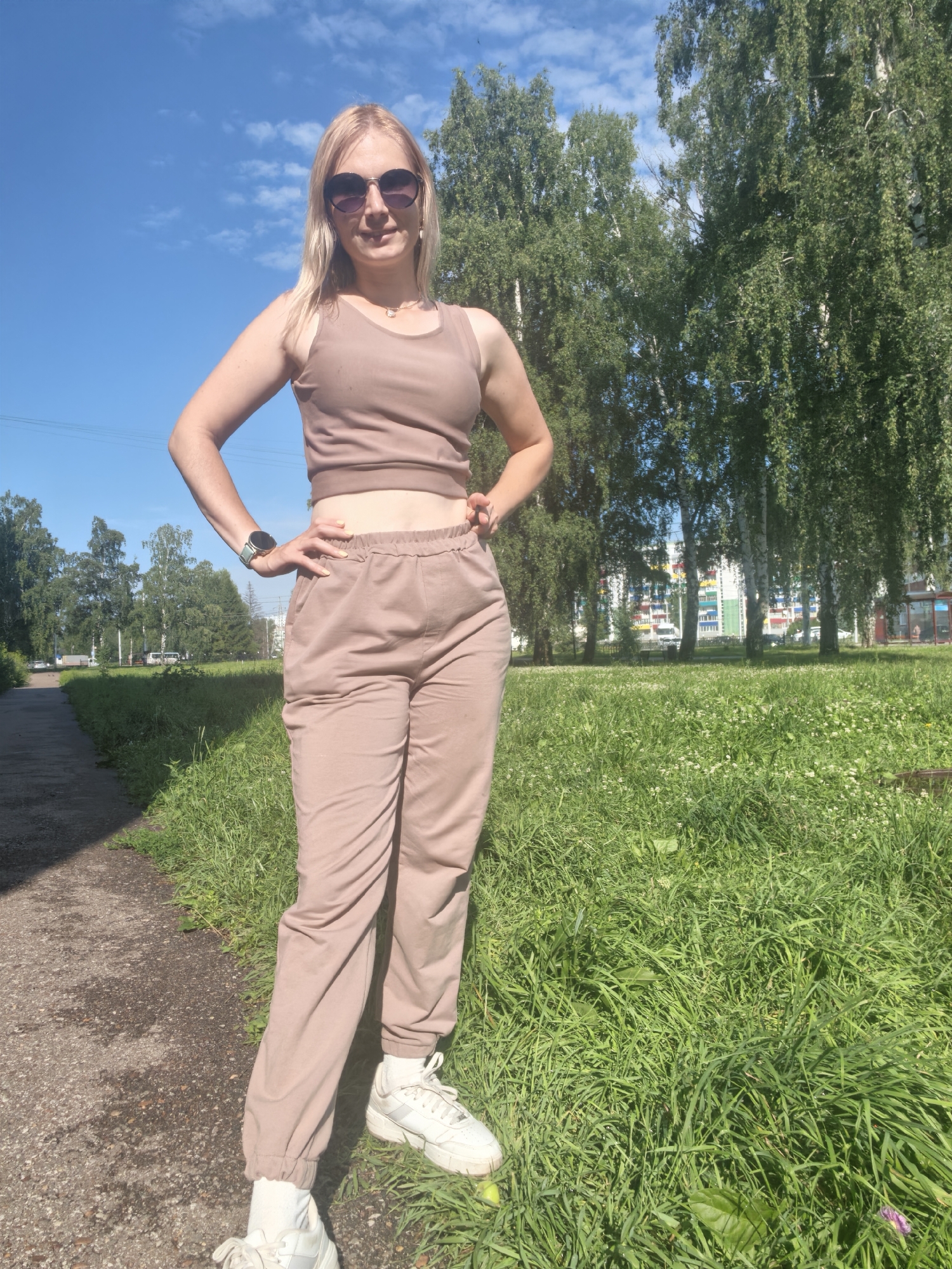 A blonde woman with sunglasses and light-colored clothing stands with her hands on her hips on a paved path next to a grassy area. Trees line the background, with a few buildings visible beyond them. The sky is clear and blue. The scene appears to be in a park or residential area.