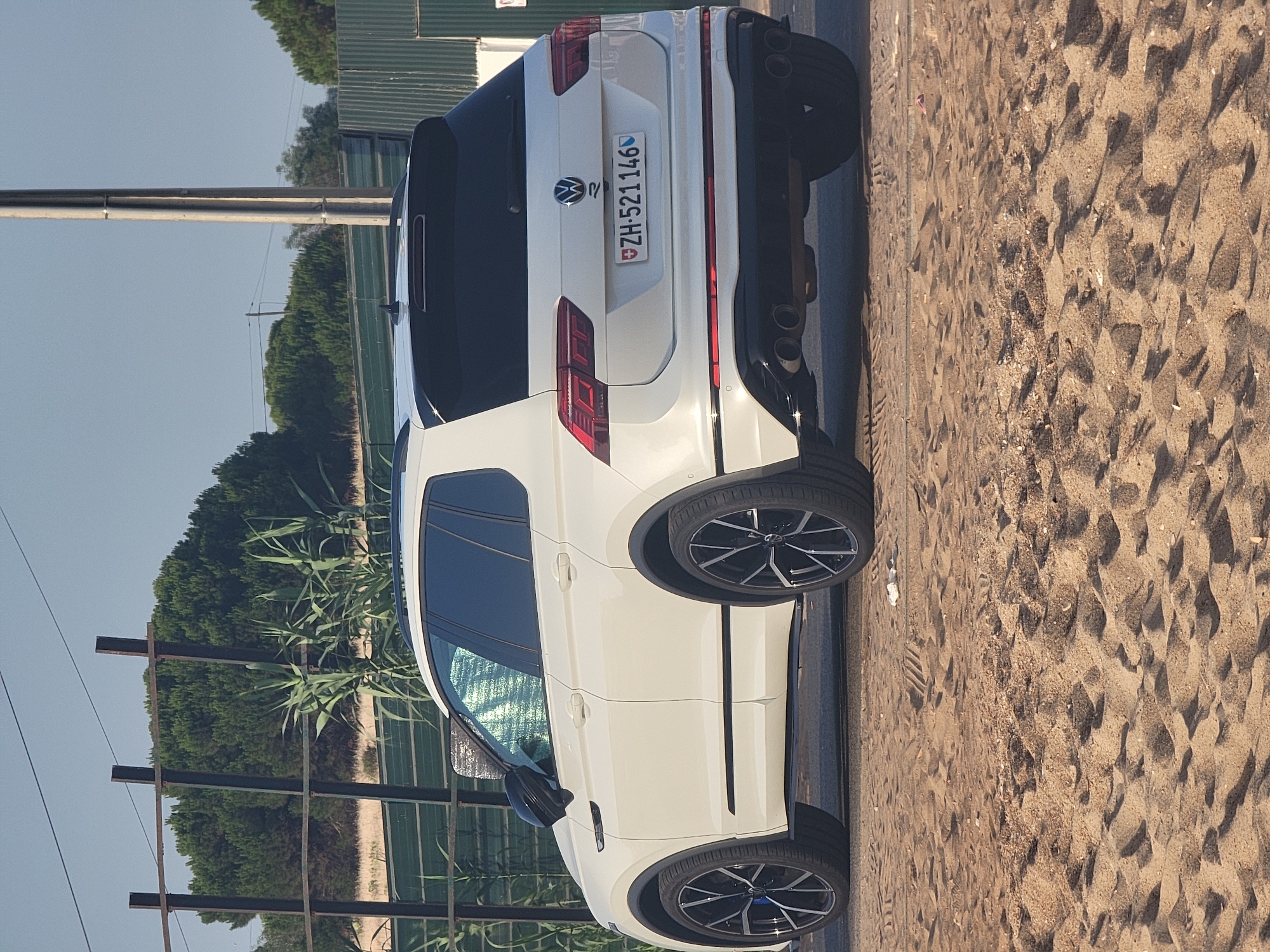 A white Volkswagen Tiguan R is parked on a sandy area next to a paved surface. The car has black trim around the wheel wells and black wheels with silver accents. The license plate is visible and reads "ZH-521146". The background shows a fence, tall green trees, and a utility pole. The scene appears to be an outdoor parking area near a beach, consistent with the coastline of Quarteira, Portugal. The sky is clear and blue.