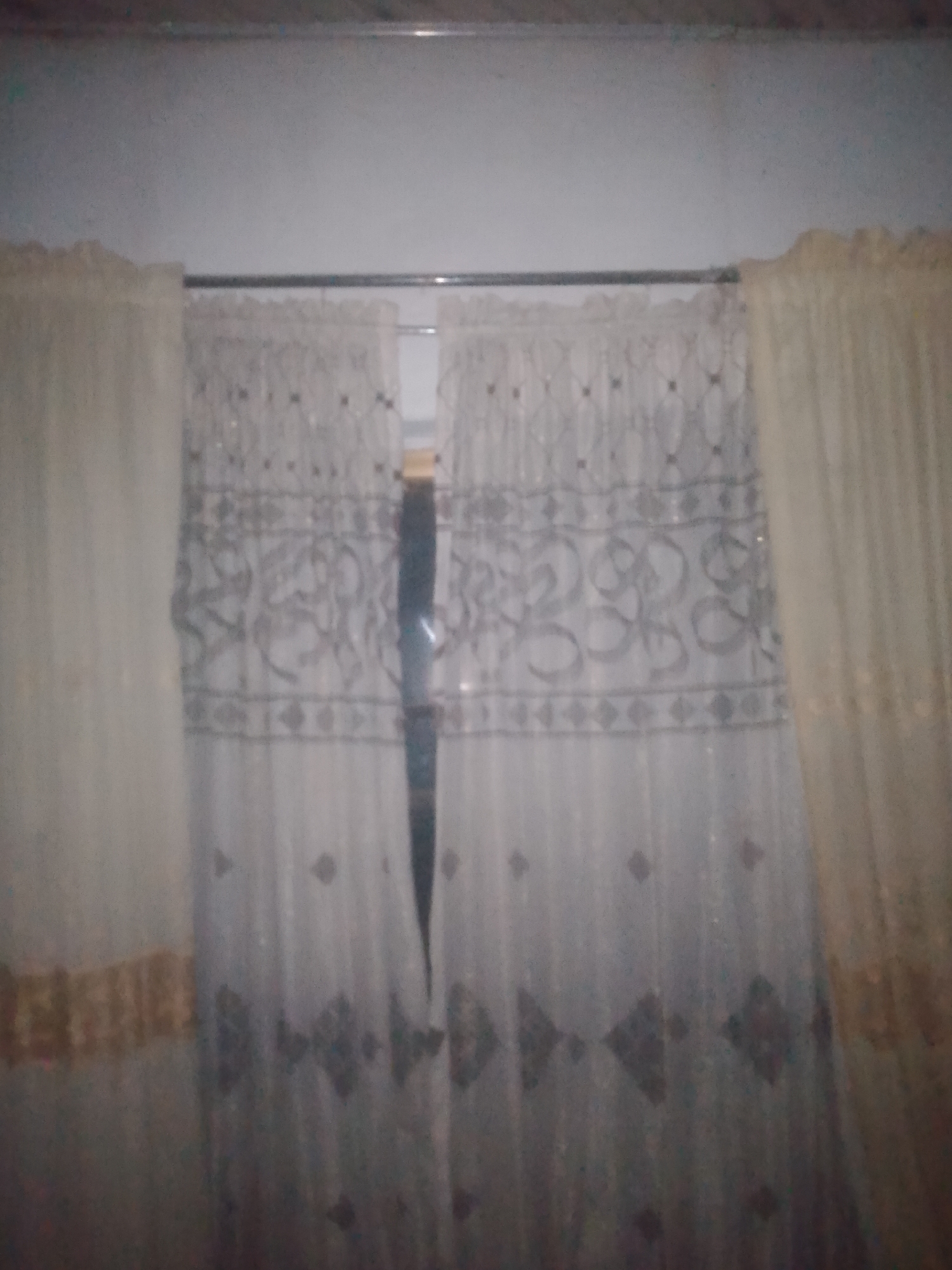 The image shows a window with cream-colored curtains on either side and a sheer, patterned curtain in the center. The sheer curtain has a horizontal band of ornate, gray swirling patterns, with smaller diamond shapes above and below it. The cream curtains appear to have a textured gold trim near the bottom. The scene is dimly lit, and the background suggests an indoor setting, likely a home in Jauro Yinu, Nigeria. The overall impression is of a decorated window, possibly in a bedroom or living area.