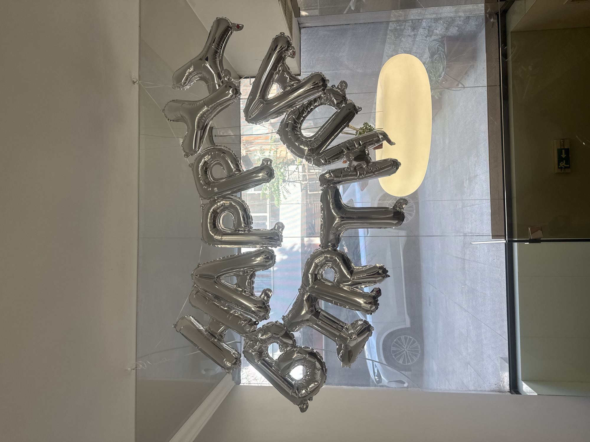 The image shows silver helium balloon letters spelling "HAPPY BIRTHDAY" suspended in front of a glass window or door. The balloons appear to be indoors, with a view to the outside through the glass. The outside scene includes a street or sidewalk, a white car, and a large, vertically oval light fixture or panel. The exact location in Varna, Bulgaria, cannot be determined from this image alone.
