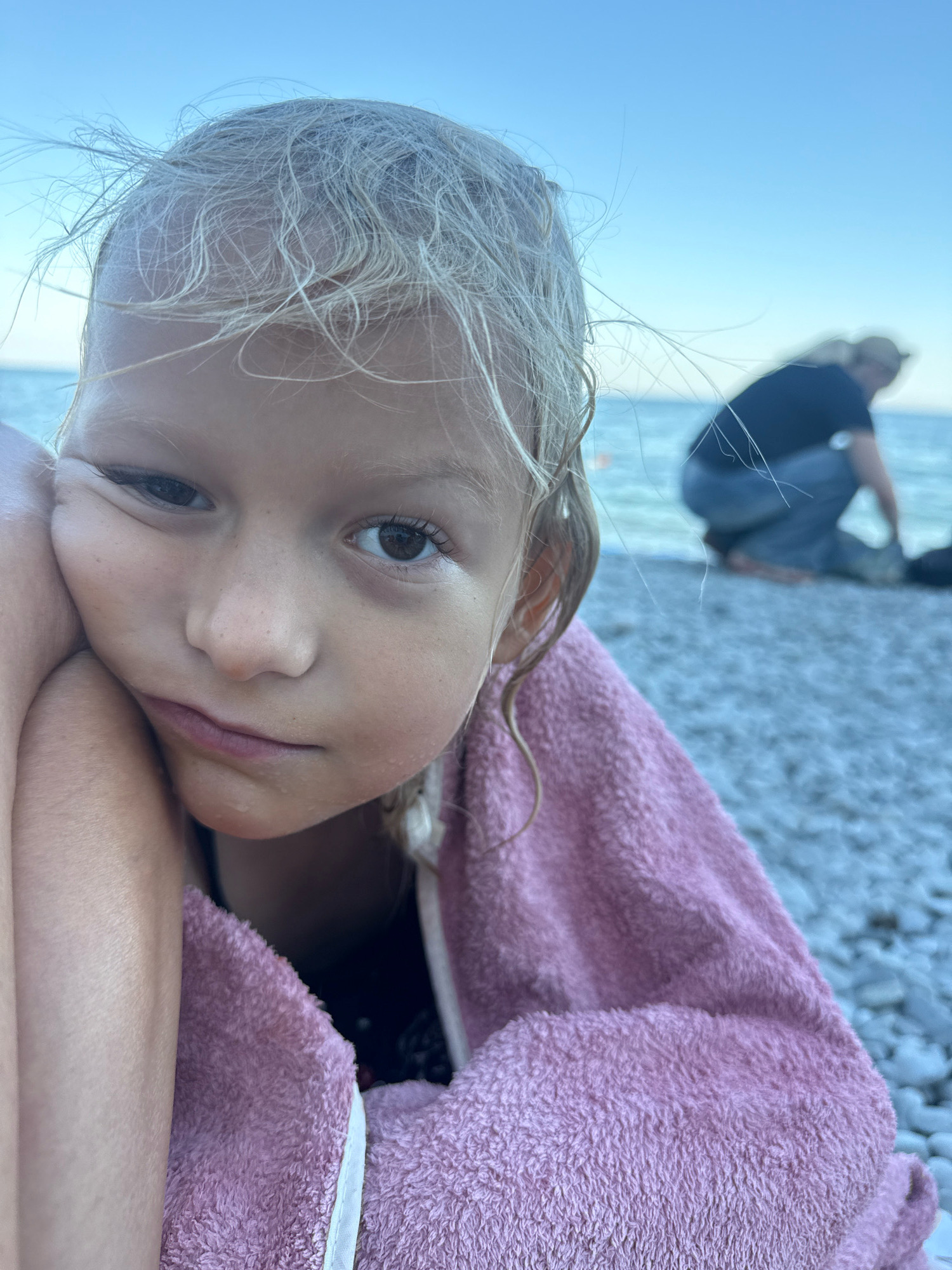 A close-up shot focuses on the face of a young child wrapped in a pink towel, resting their head on their arm. The child has blonde hair and is looking directly at the camera with a neutral expression. In the background, a person is crouched near the water on a pebble beach, with the sea and a clear sky visible. The location is identified as Partenit, Ukraine.