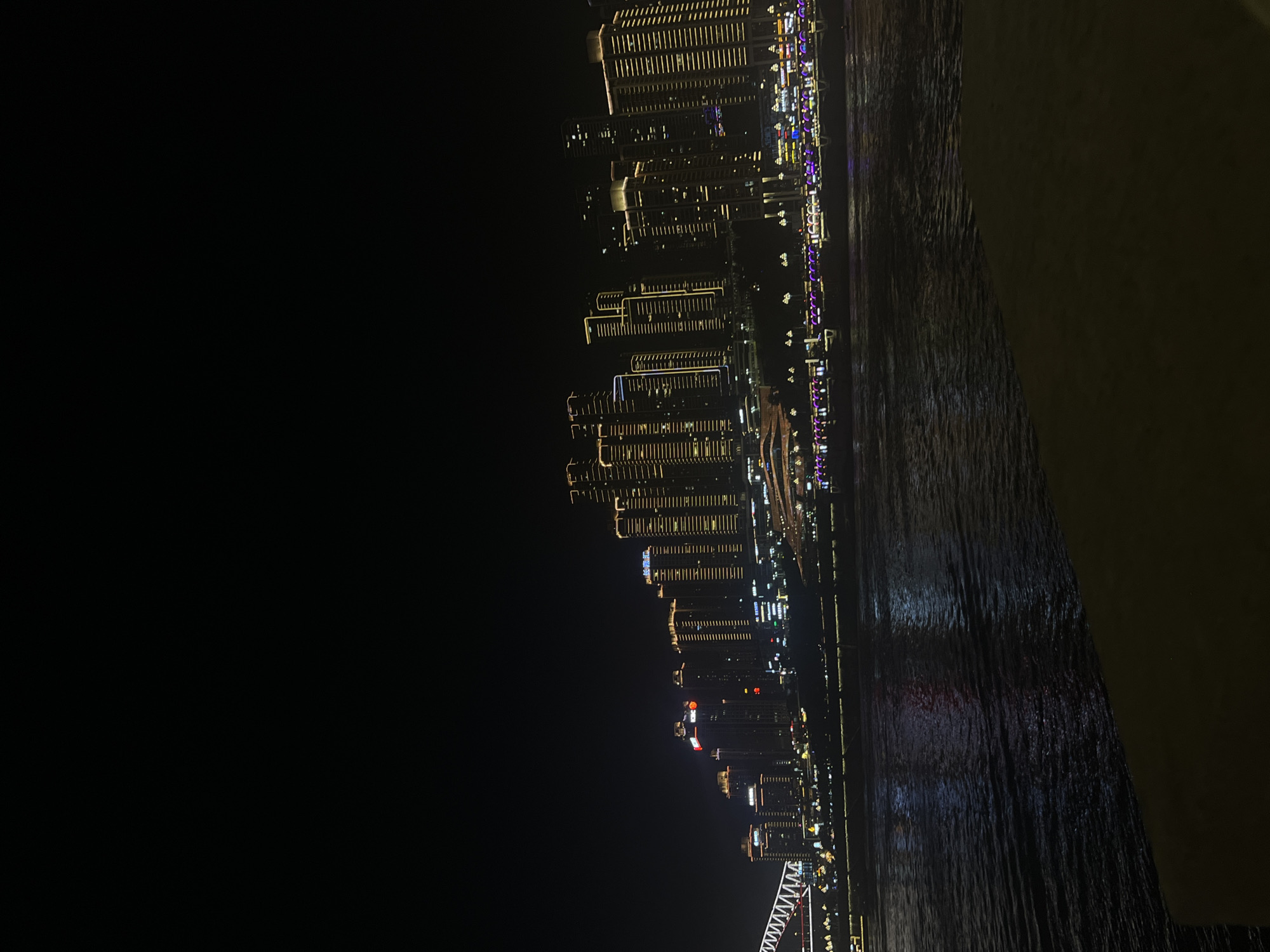 This is a nighttime, wide-angle, aerial shot of a cityscape with a river. The image is oriented vertically, with the cityscape on the left and the river on the right, suggesting the photo was taken from a high vantage point overlooking the city from across the water. The city skyline is illuminated by countless lights from skyscrapers, creating a dazzling spectacle. The buildings are a mix of modern high-rises, many with horizontal and vertical lines of light that highlight their structures. Some buildings have colorful lights, including purples, blues, and reds, contributing to the vibrant urban panorama. The river reflects the city lights, creating shimmering patterns on its dark surface. The water appears choppy, with visible ripples. The sky is dark, indicating it is nighttime, and the weather seems clear. The overall mood is one of urban grandeur and nighttime beauty, with the city's lights creating a sense of energy and dynamism. The scene is set in the Yuzhong District of China. No people are discernible in the image, but the focus is on the impressive architectural landscape.